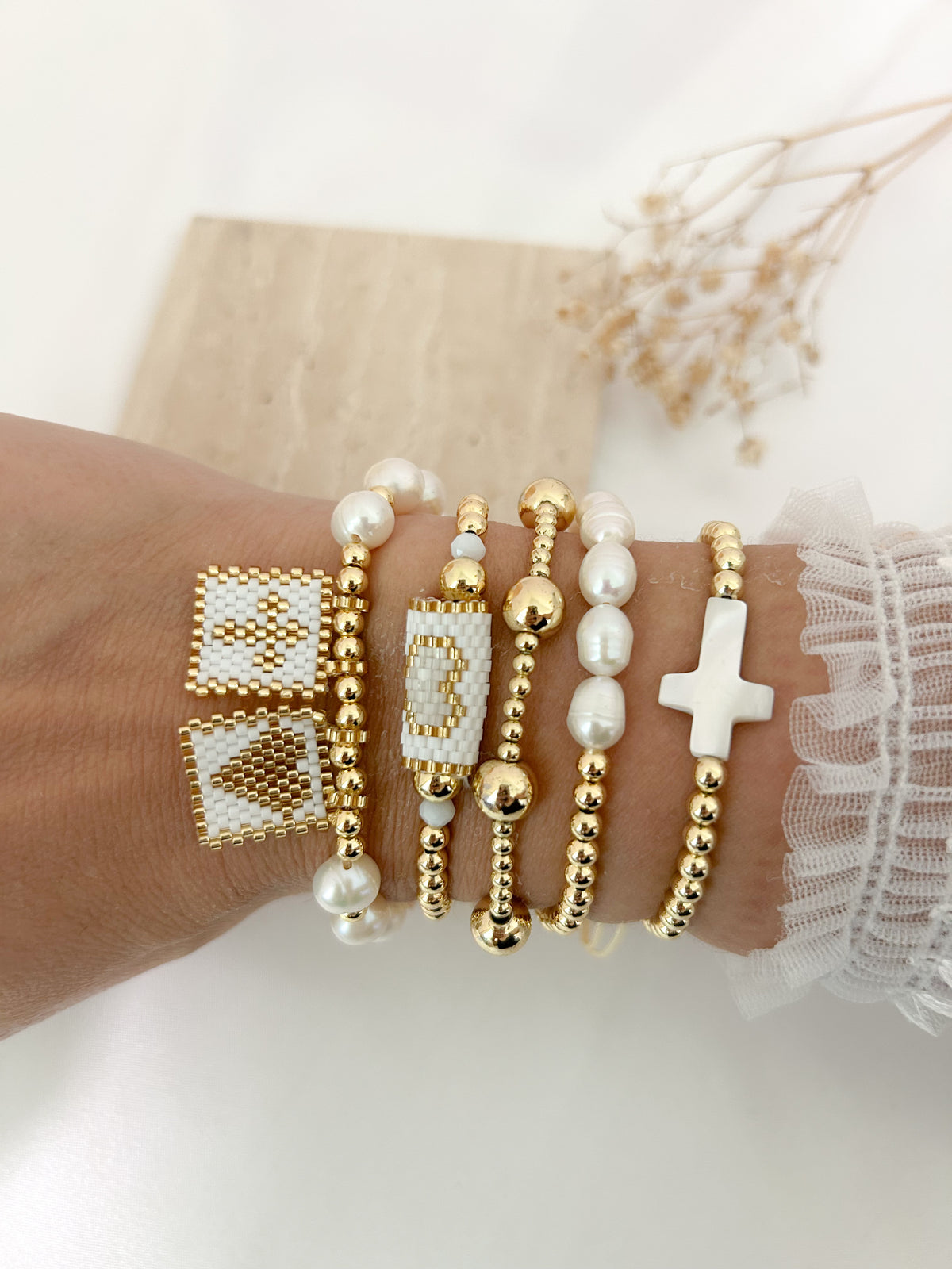 A close-up of a person's wrist wearing multiple delicate bracelets, including gold beaded strands, white pearl-like beads, a white cross charm, and two square beaded bracelets with gold and white detailing.