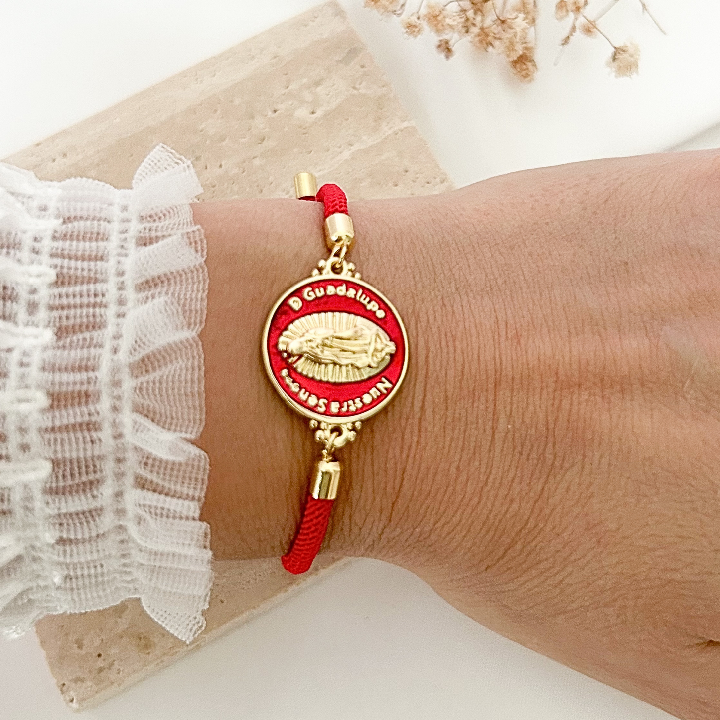 Adjustable red cord bracelet featuring a round Virgin Mary medal with gold-tone details and vibrant red enamel accents, styled on a neutral stone surface with soft dried florals.