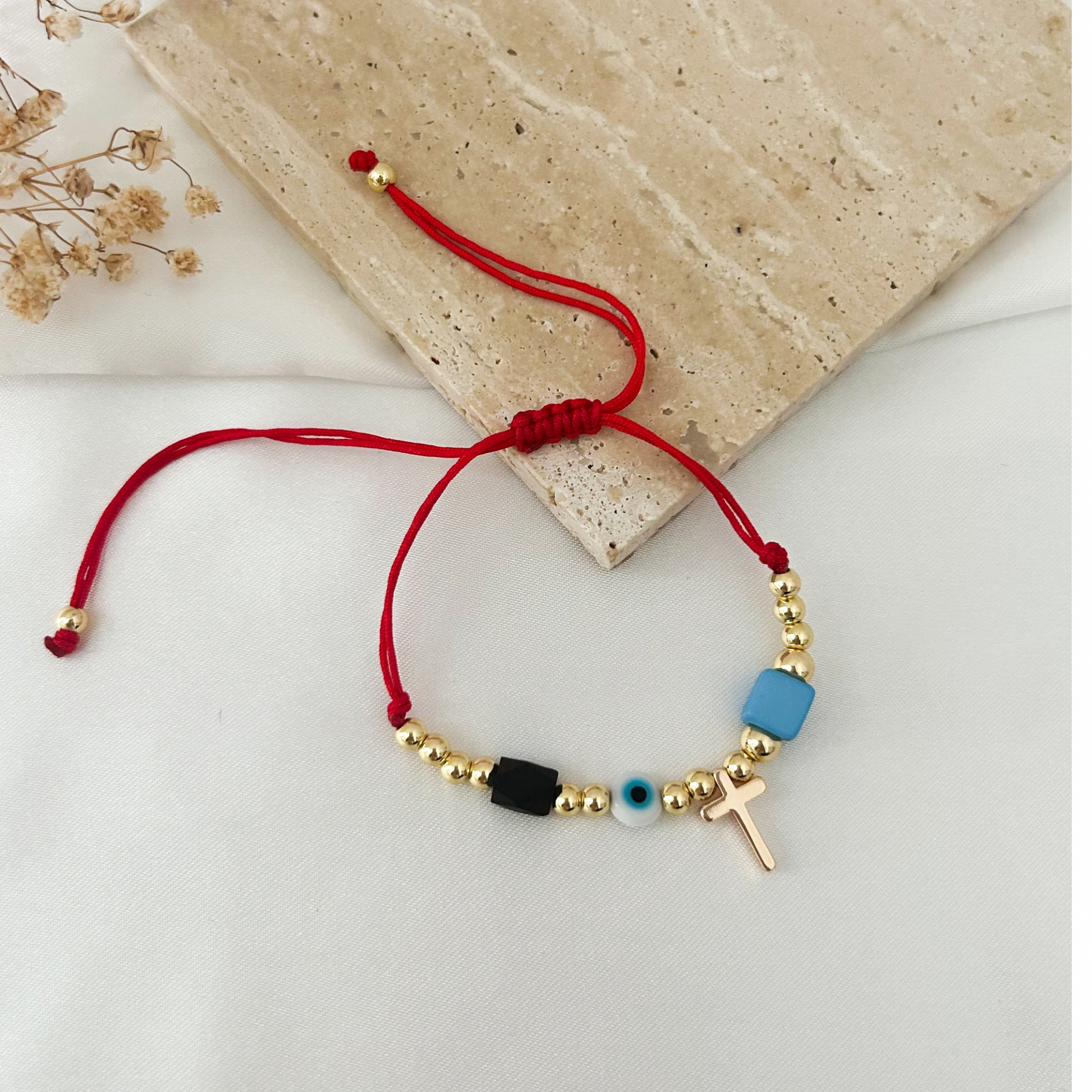 Minimalist adjustable bracelet with a vibrant red cord, gold-tone beads, a gold cross charm, a blue evil eye centerpiece, and contrasting black and blue beads. A stylish symbol of faith and protection with a contemporary design.