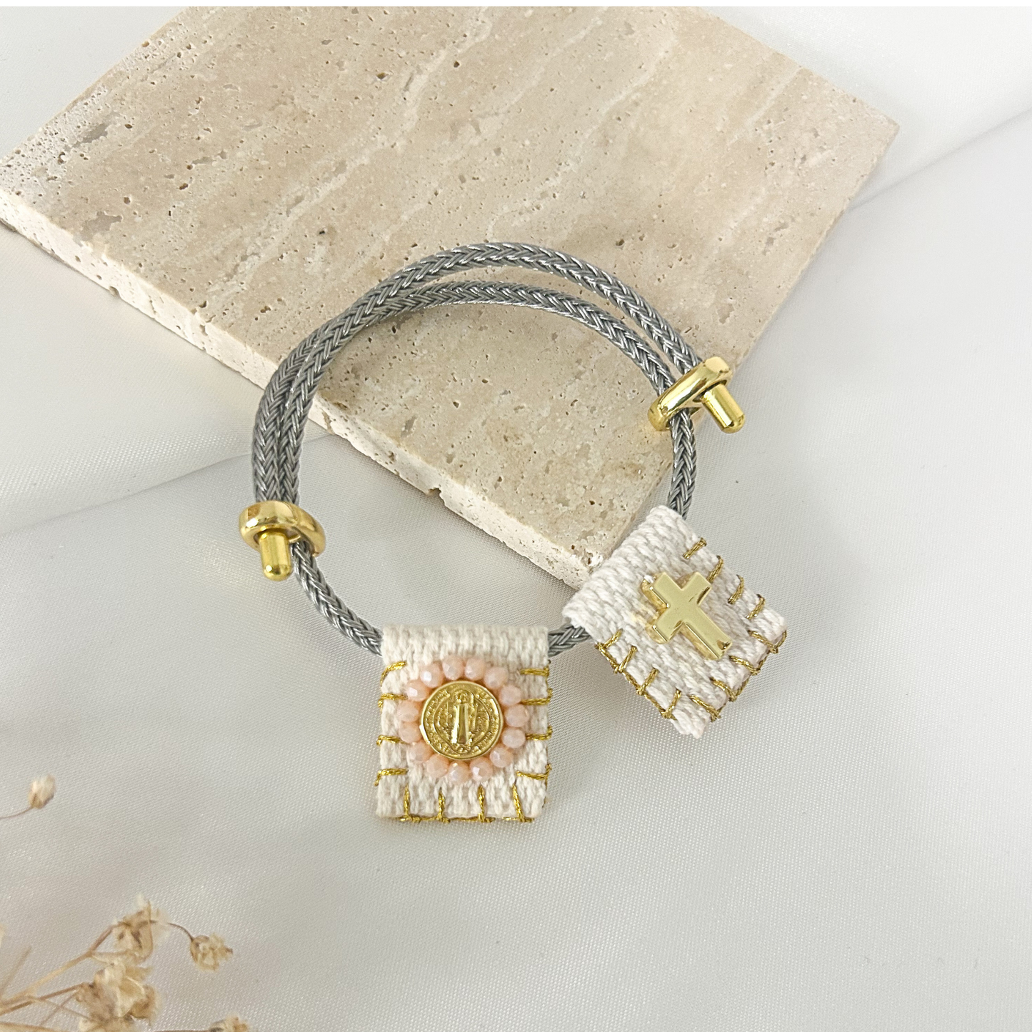 A double-strand gray braided cord bracelet with gold-toned hardware, featuring two woven rectangular charms: one white with a gold cross and one white with a gold medallion, displayed on a light stone slab.
