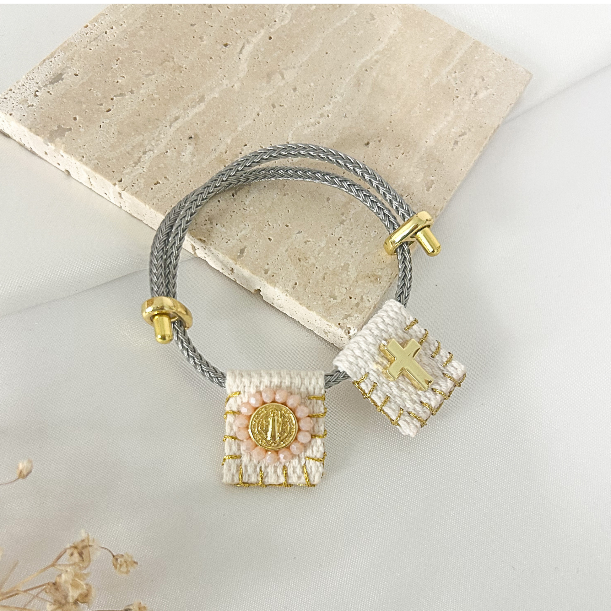 A double-strand gray braided cord bracelet with gold-toned hardware, featuring two woven rectangular charms: one white with a gold cross and one white with a gold medallion, displayed on a light stone slab.