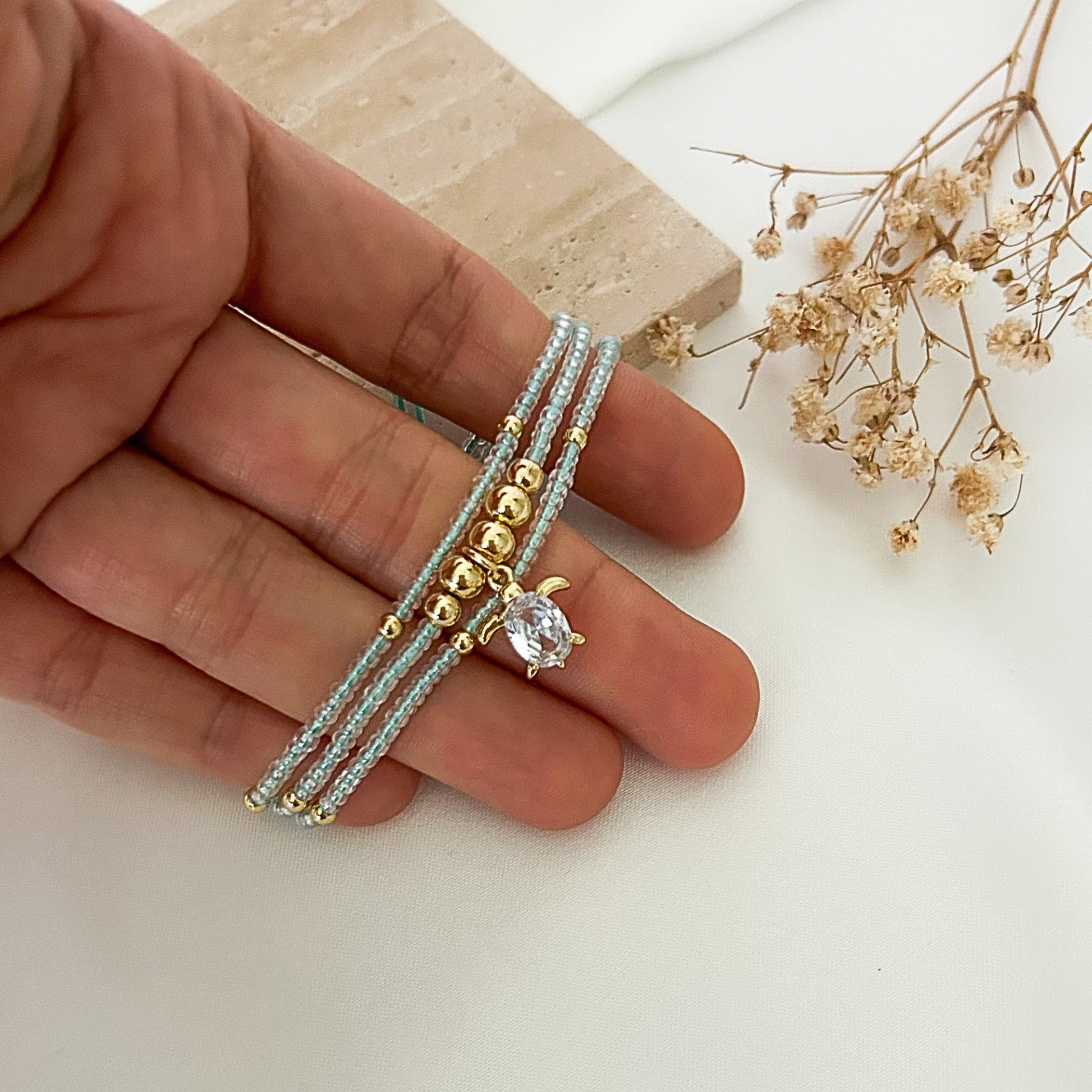 Close-up of a multi-strand aqua beaded bracelet with gold-tone beads and a shimmering turtle pendant, photographed in hand with dried flowers and a natural stone prop.