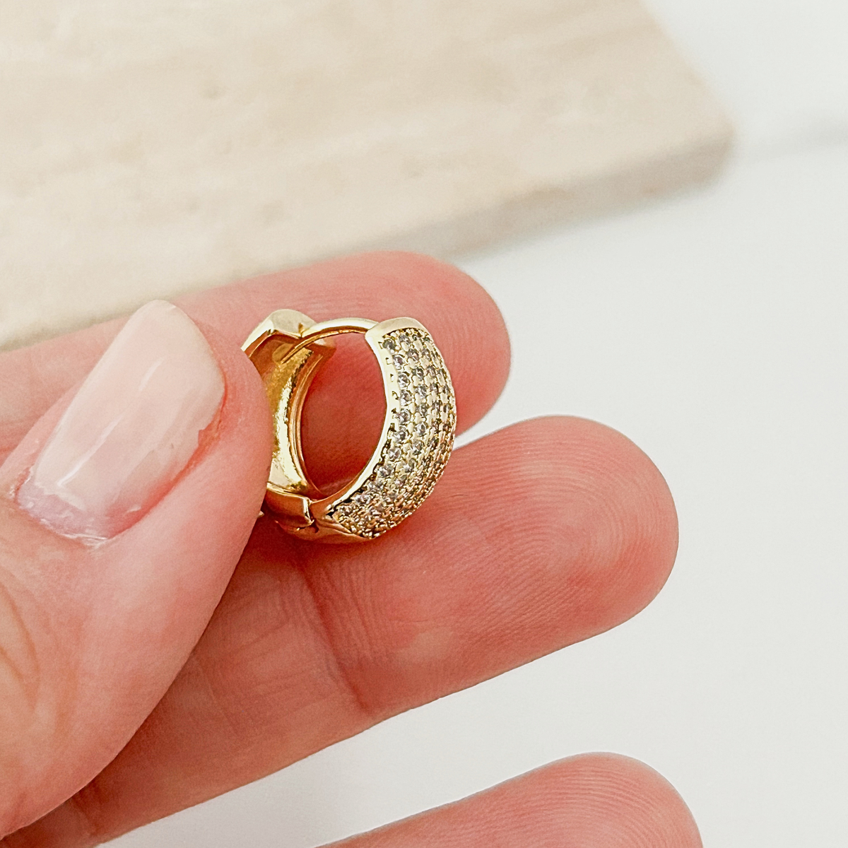 A close-up of a hand holding a small gold huggie hoop earring paved with clear stones, with a light beige stone slab in the background.