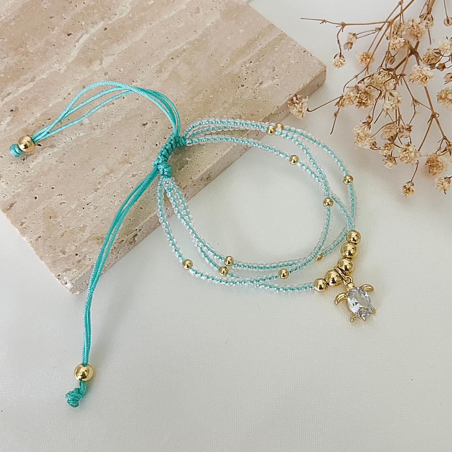 Delicate adjustable bracelet featuring three strands of light turquoise seed beads, gold-tone accents, and a sparkling turtle charm with a clear crystal centerpiece, displayed on a neutral stone surface.