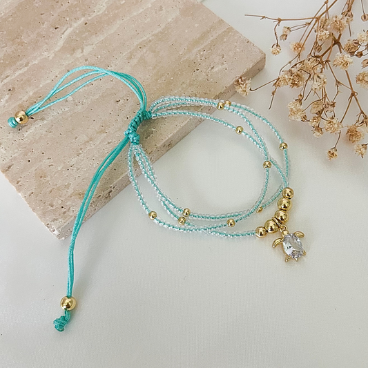 Delicate adjustable bracelet featuring three strands of light turquoise seed beads, gold-tone accents, and a sparkling turtle charm with a clear crystal centerpiece, displayed on a neutral stone surface.