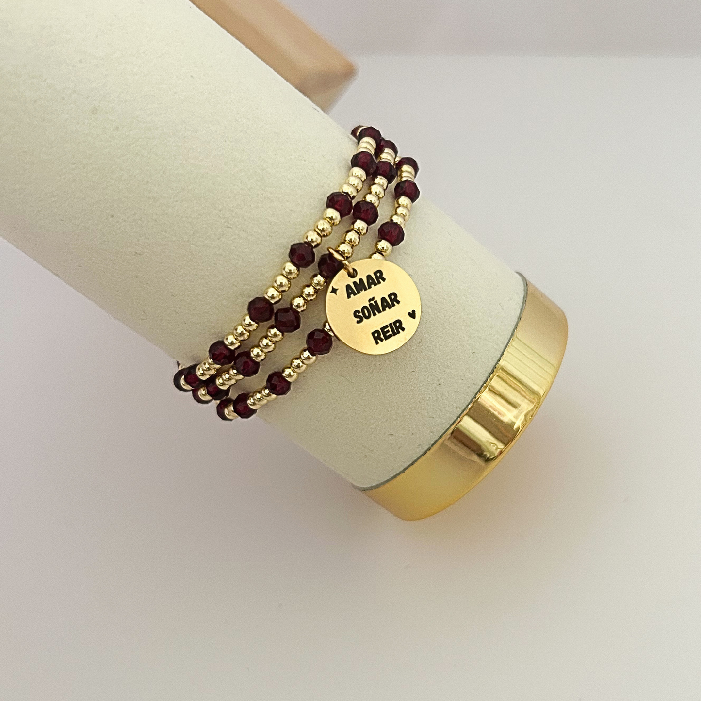 Burgundy and gold beaded wrap bracelet with engraved “Amar Soñar Reír” charm on jewelry display.