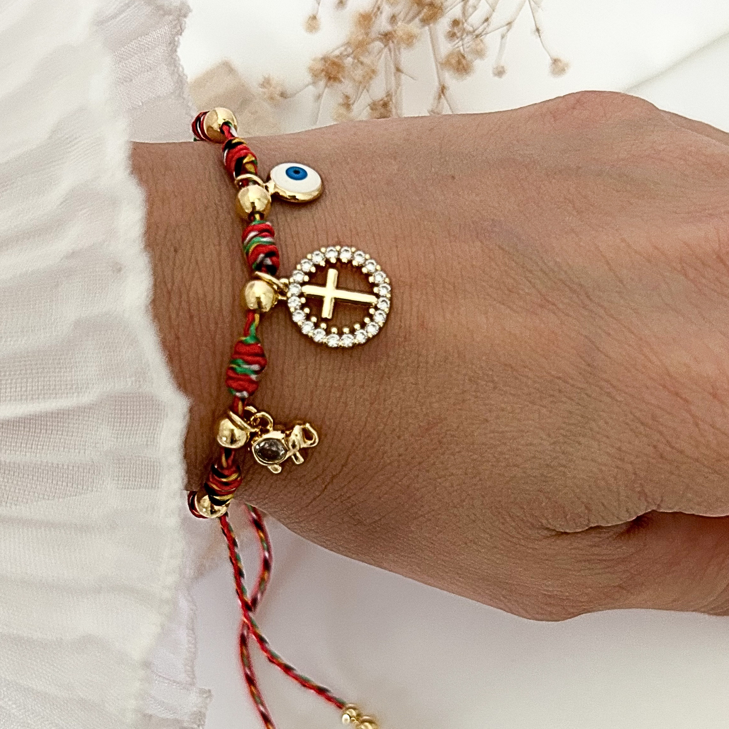 Adjustable braided bracelet in red, green, black, and white tones featuring gold-tone accents, a sparkling cross charm, an evil eye charm, and a small elephant charm, worn on the wrist.