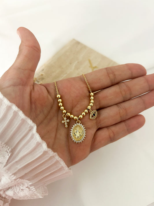 A gold-toned beaded necklace with a large oval religious pendant, a small cross charm, and a dark oval charm, displayed on an open hand.