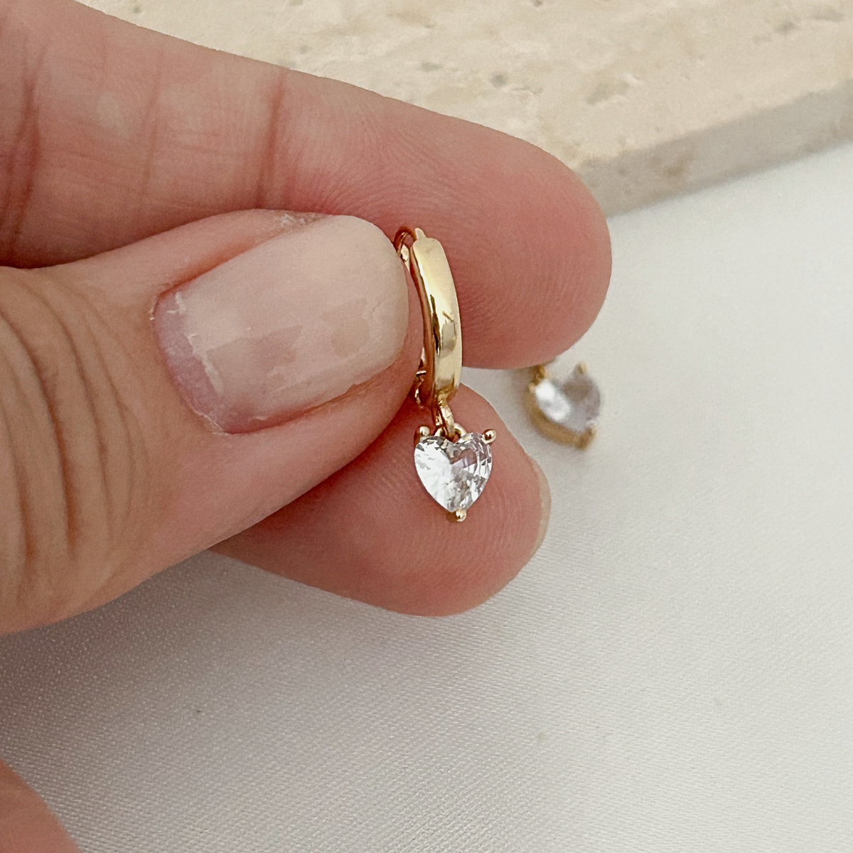 A close-up of a hand holding a small gold huggie hoop earring with a dangling heart-shaped clear gemstone, with another matching earring resting nearby on a light surface.