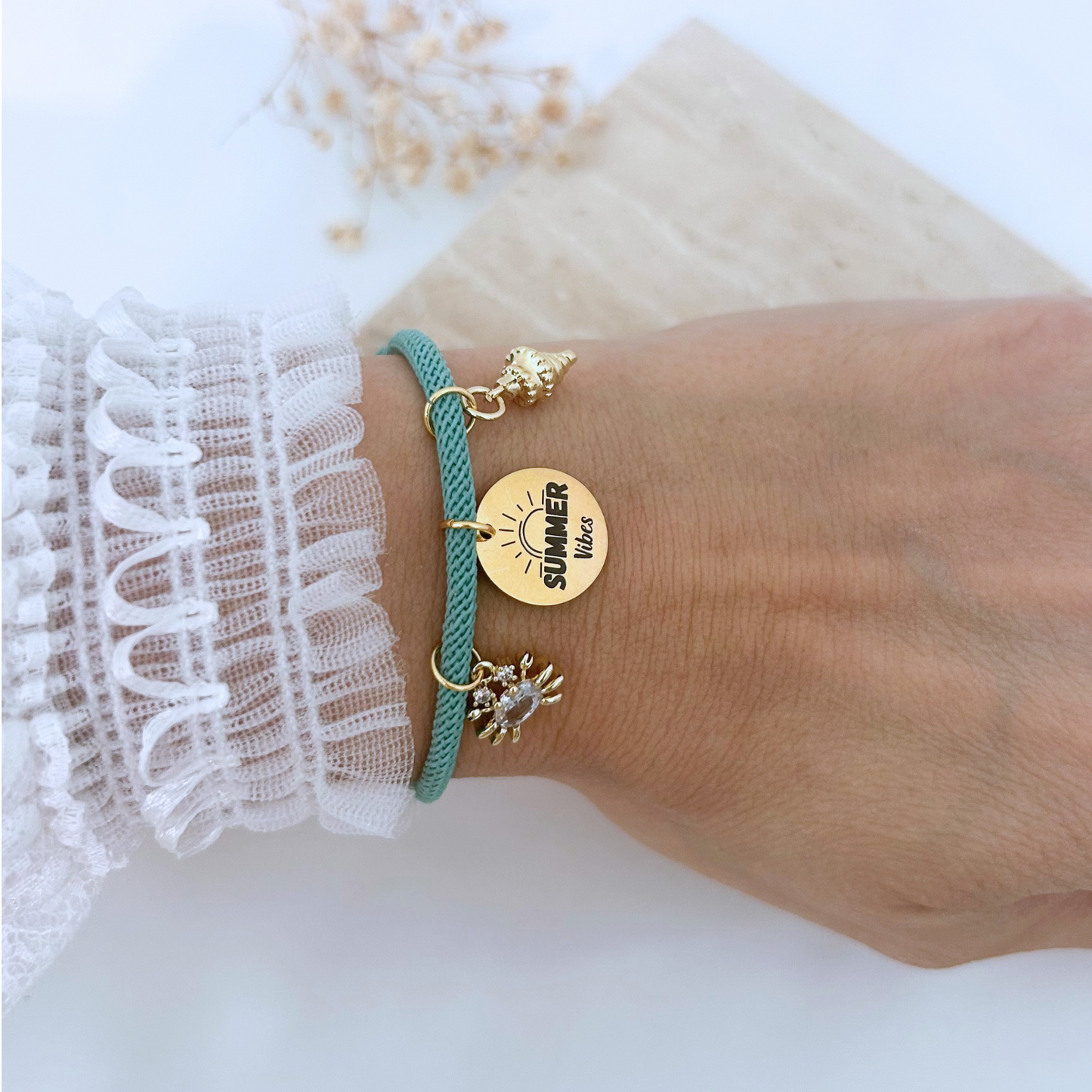 Close-up of a teal adjustable bracelet worn on the wrist, showcasing gold beach-themed charms including a seashell, a crystal crab, and a “Summer Vibes” pendant. A fun and playful accessory for sunny days.
