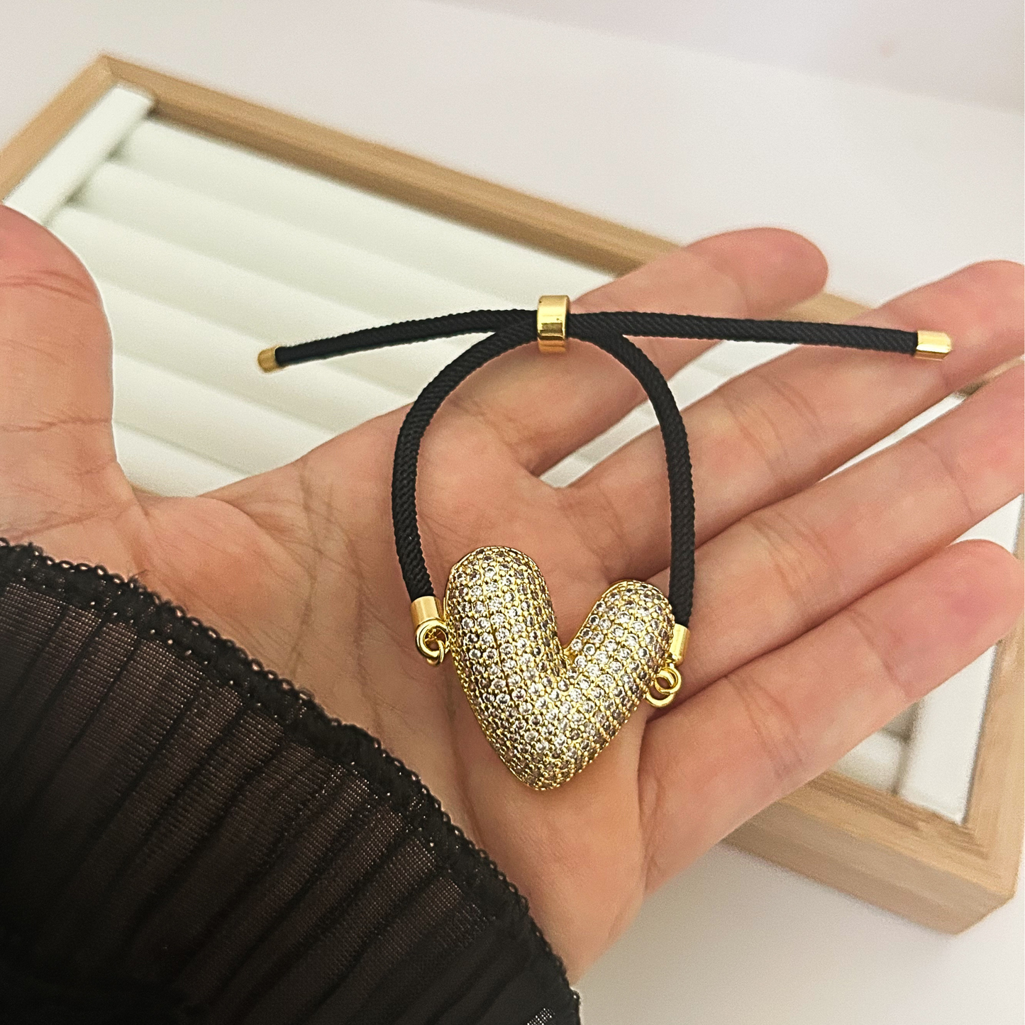 Hand holding gold heart bracelet with sparkling crystal design and black cord.