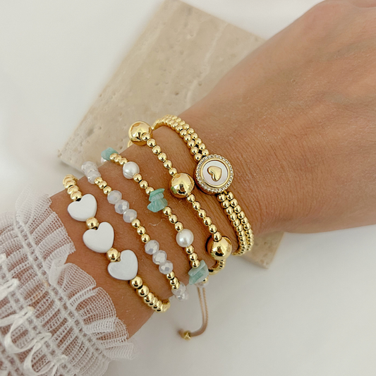 Gold bracelet stack featuring white hearts, crystal beads, turquoise accents, pearls, and a sparkling heart medallion.