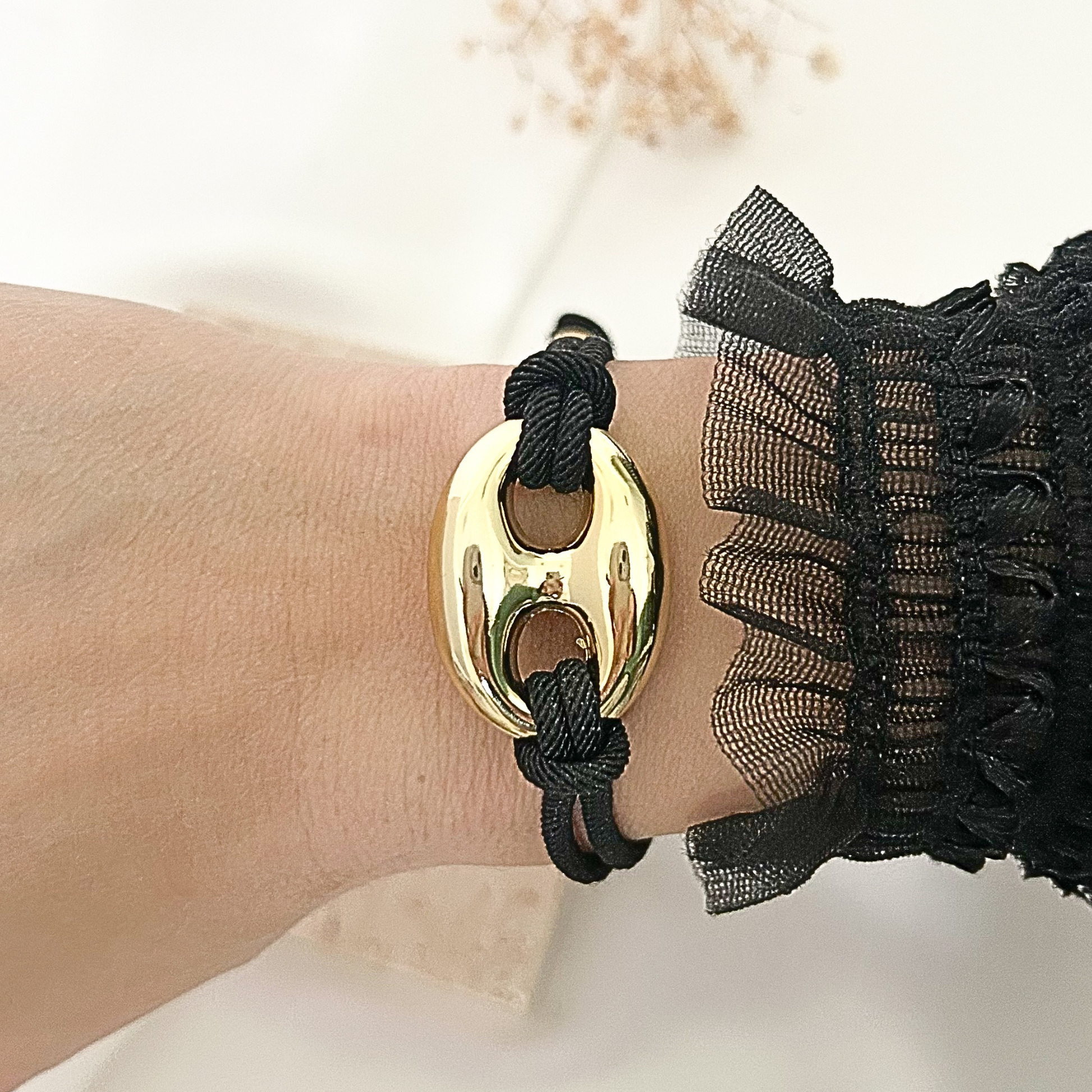 Black adjustable cord bracelet featuring a bold gold-tone oval centerpiece, styled on the wrist with a sheer black ruffle sleeve against a soft neutral background.