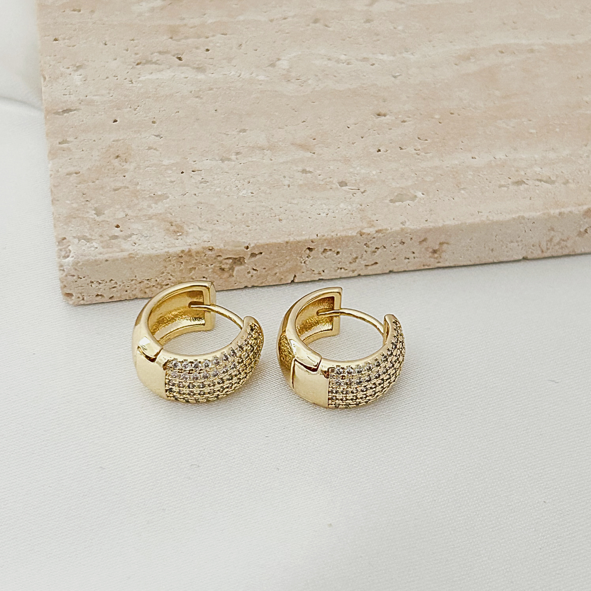 A pair of gold huggie hoop earrings with textured pavé detailing, displayed on a white surface with a beige stone tile in the background.