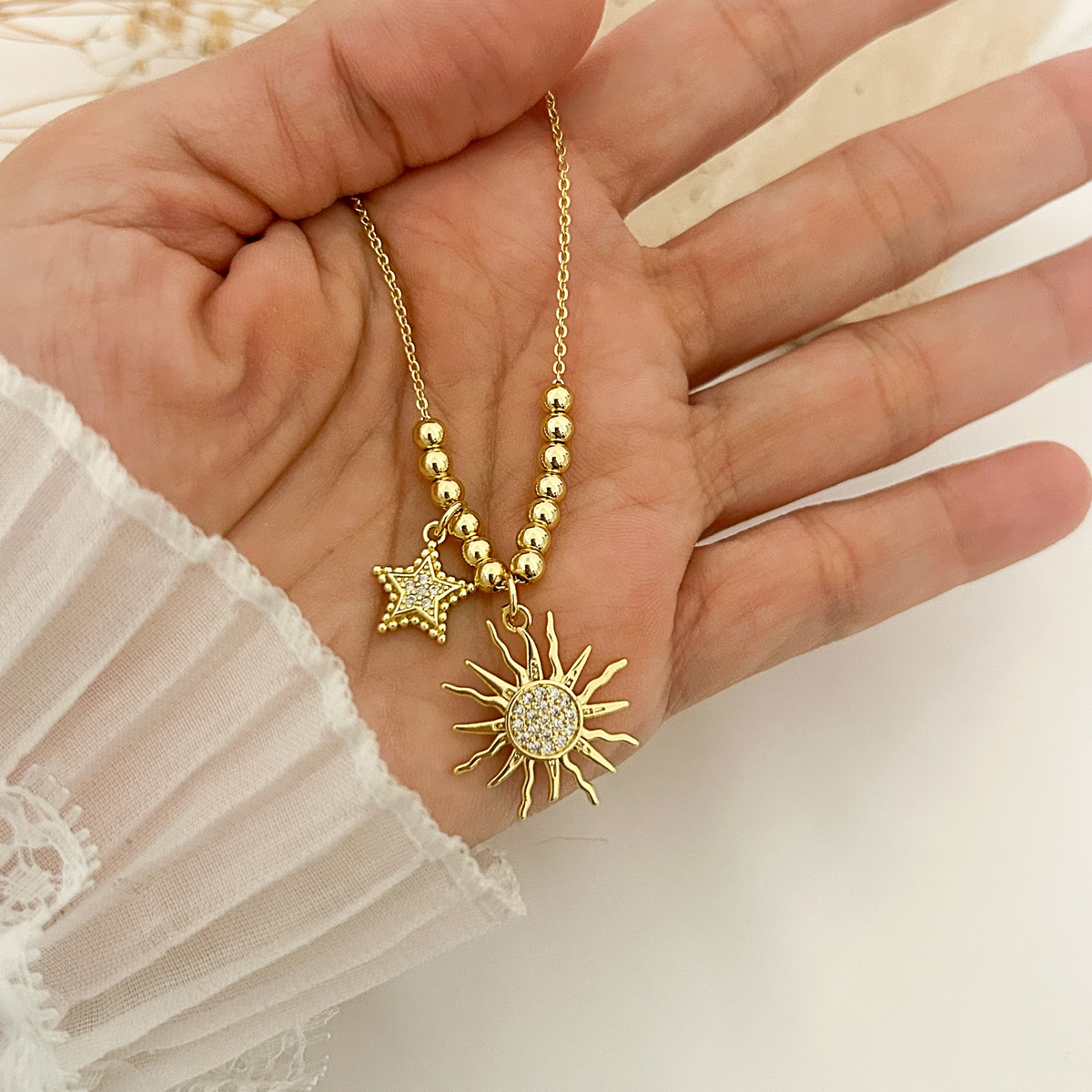 A gold-toned necklace with a beaded chain, a star charm, and a sunburst pendant with a crystal-encrusted center, held in a person's hand.