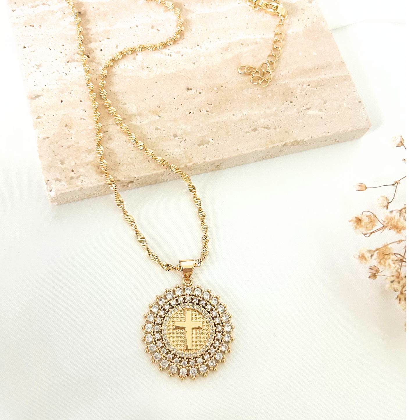 Gold-tone necklace featuring a round crystal-studded pendant with a cross centerpiece, displayed on a neutral stone background with delicate dried florals. A timeless faith-inspired statement piece.