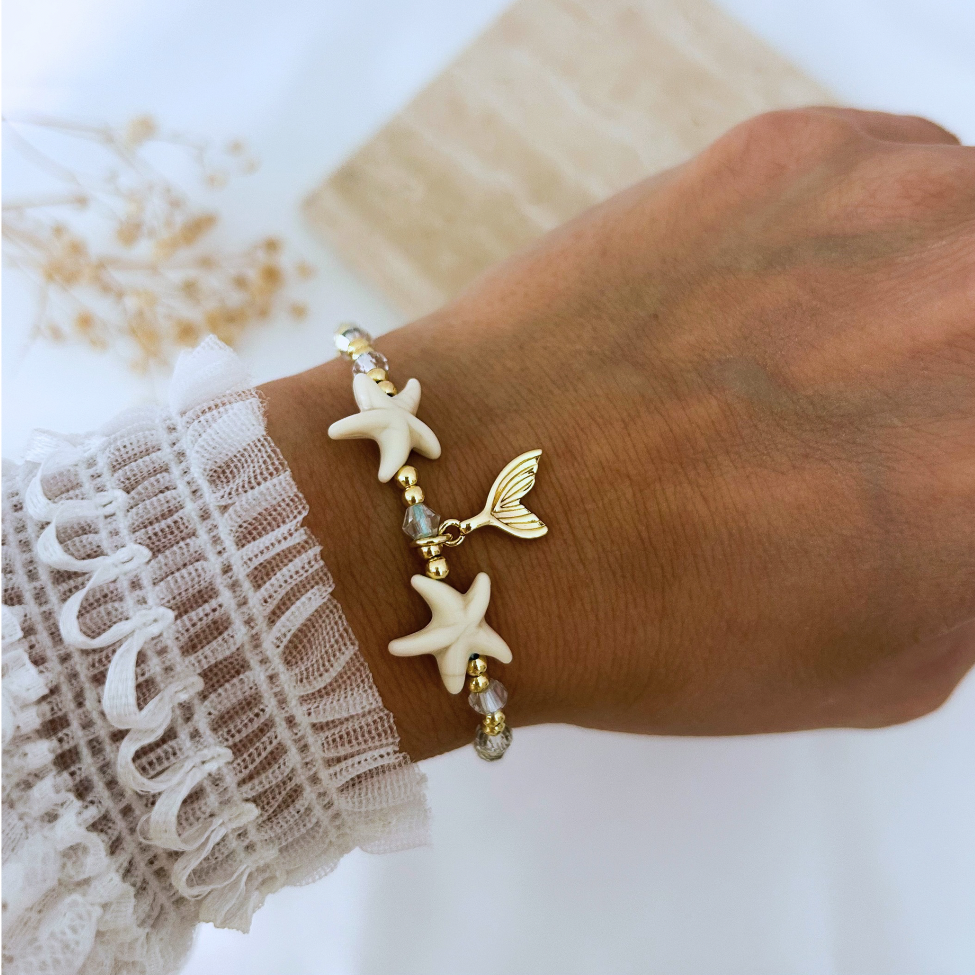 Adjustable beach-inspired bracelet worn on the wrist, featuring white starfish beads, shimmering crystal accents, polished gold beads, and a delicate gold mermaid tail charm. A fresh coastal accessory perfect for summer