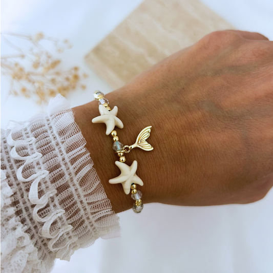 Adjustable beach-inspired bracelet worn on the wrist, featuring white starfish beads, shimmering crystal accents, polished gold beads, and a delicate gold mermaid tail charm. A fresh coastal accessory perfect for summer