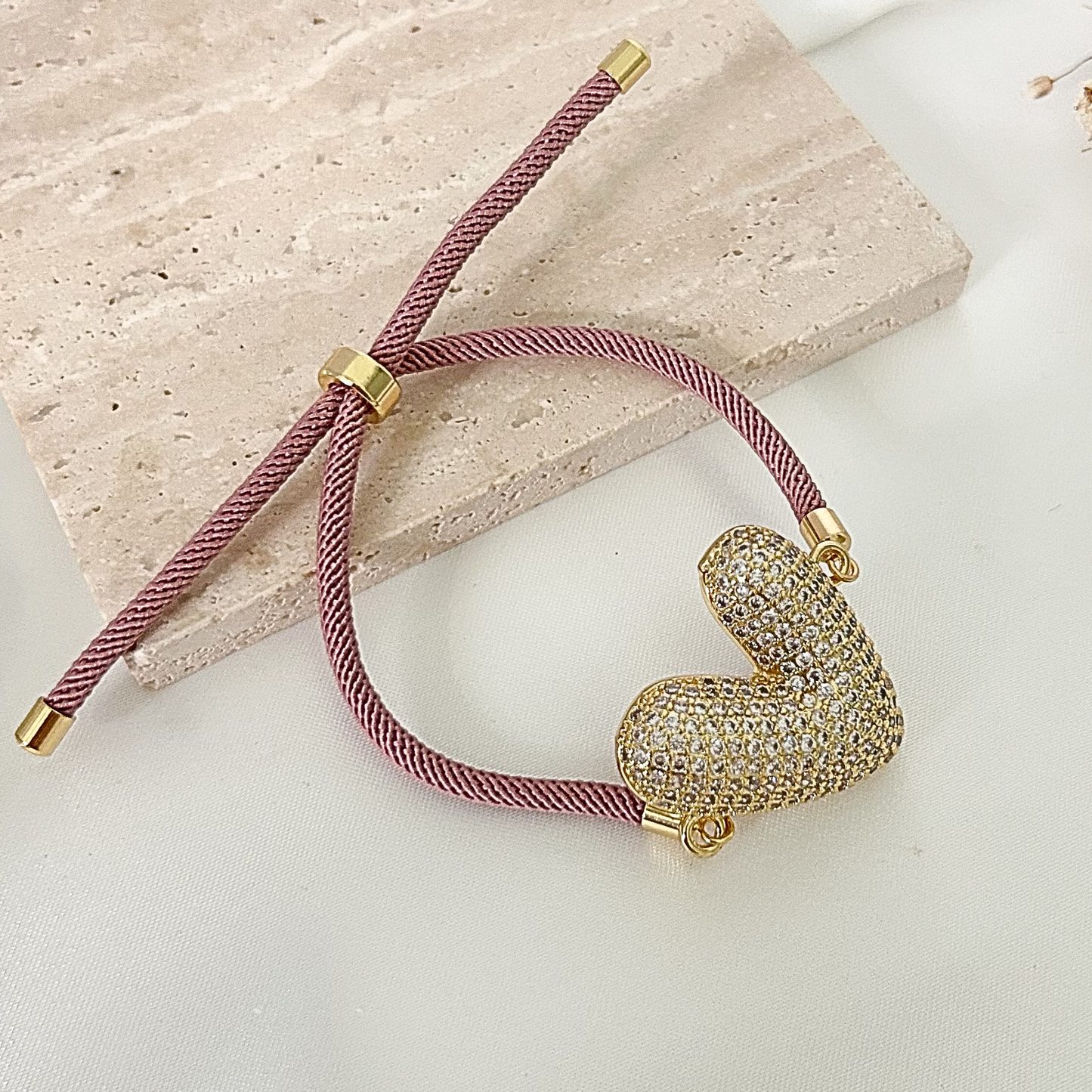 Elegant heart bracelet with a pavé crystal heart centerpiece, dusty rose adjustable cord, and gold-tone details displayed on a neutral background.