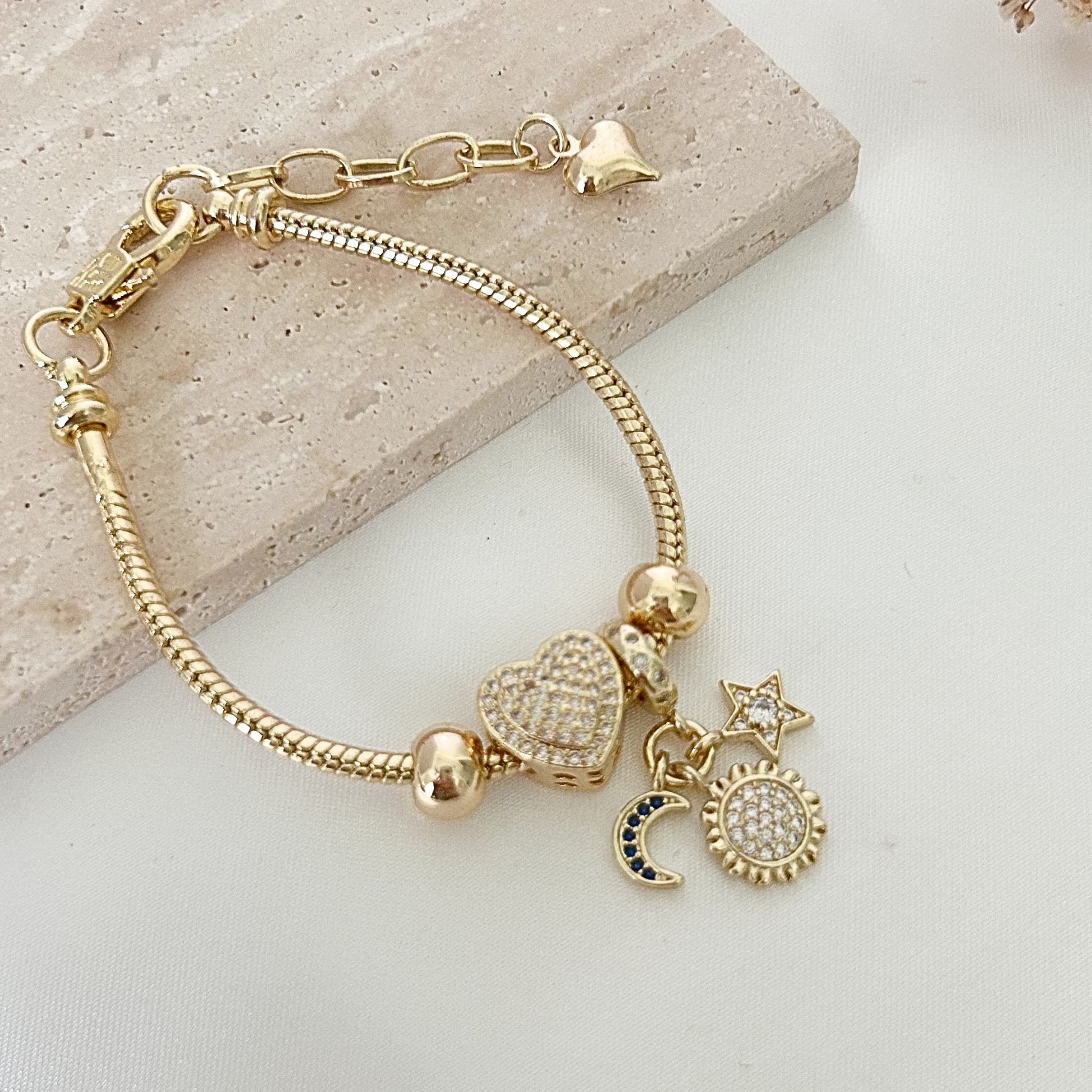Gold-tone charm bracelet displayed on a neutral stone surface, featuring a sparkling heart centerpiece and celestial moon, sun, and star charms adorned with crystal accents.
