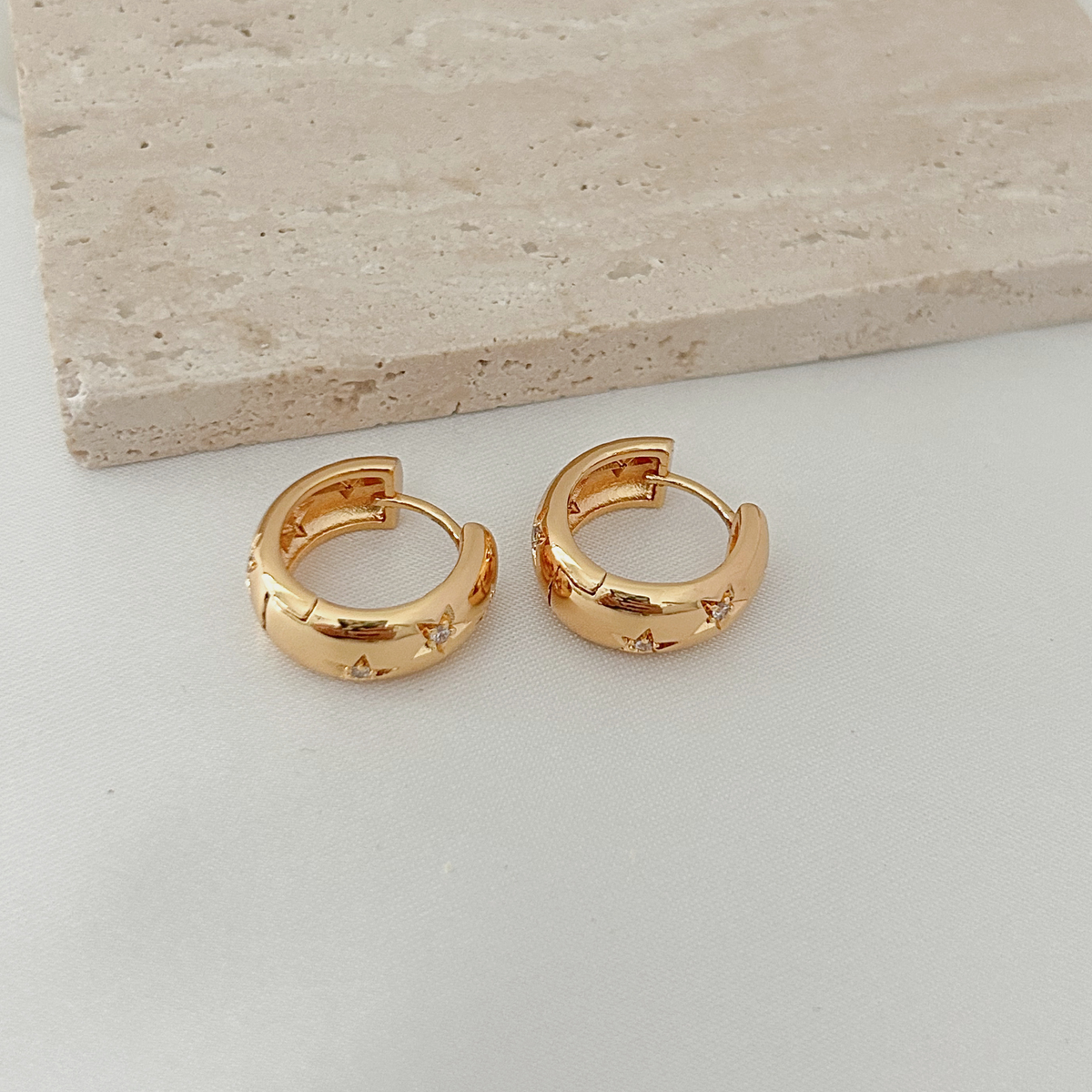 A pair of gold huggie hoop earrings with small star cutouts, displayed on a light gray surface next to a beige stone block.