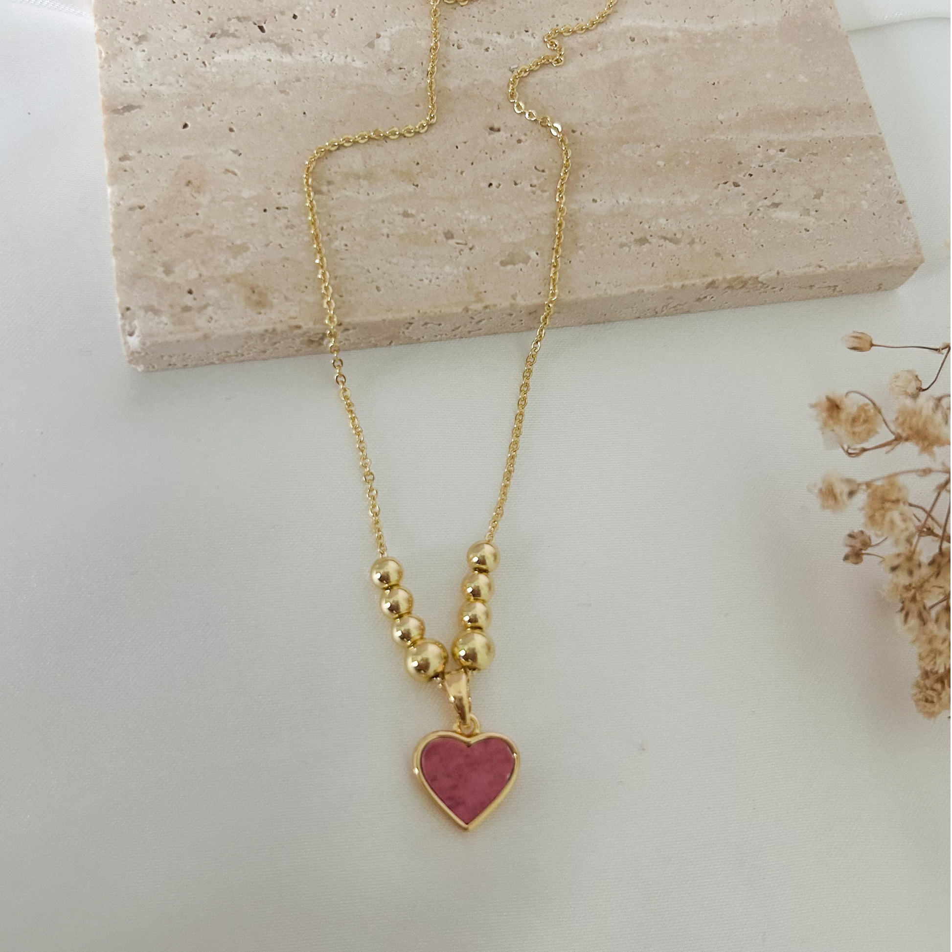 Close-up of a gold necklace with a rosy pink heart charm framed in gold and complemented by graduated gold bead accents for an elegant and romantic touch.