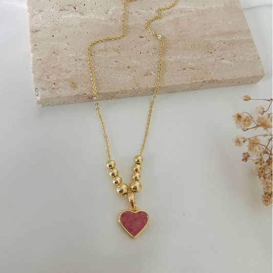 Close-up of a gold necklace with a rosy pink heart charm framed in gold and complemented by graduated gold bead accents for an elegant and romantic touch.