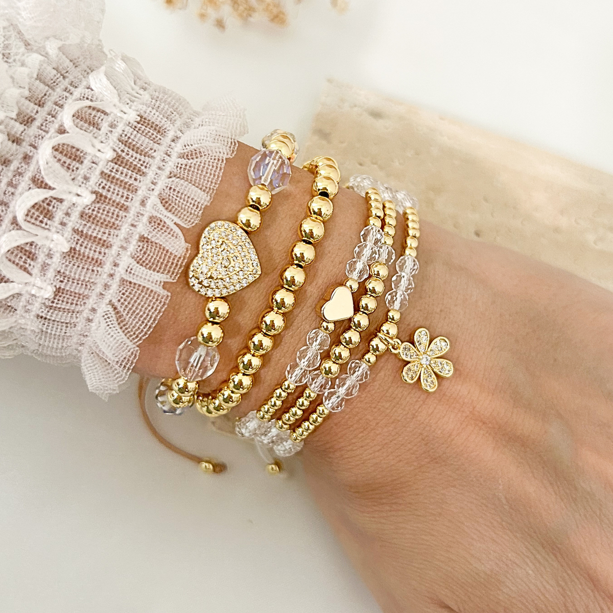 Gold and crystal bracelet stack featuring pavé heart, gold bead bracelets, faceted crystal beads, a heart accent, and a sparkling flower charm. Elegant everyday jewelry for layering or wearing individually.