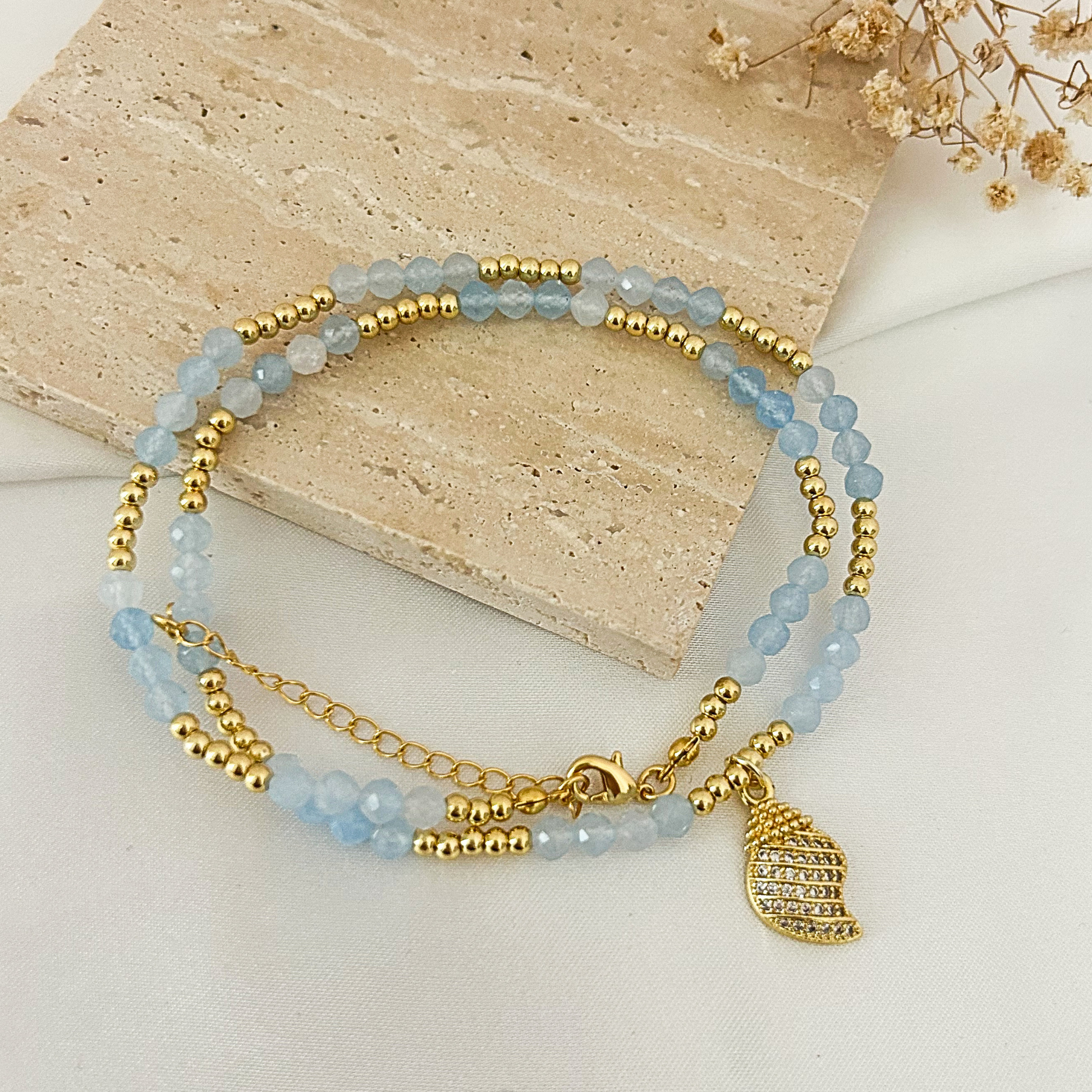 A double-strand beaded bracelet with light blue faceted beads, gold spacer beads, a gold chain extension, and a gold leaf-shaped charm embellished with clear rhinestones, displayed on a beige stone slab.