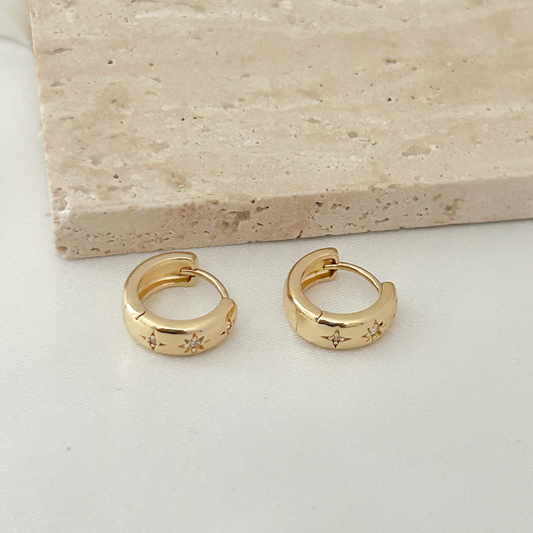 A pair of gold huggie hoop earrings with small star details, displayed on a light surface next to a beige stone tile.