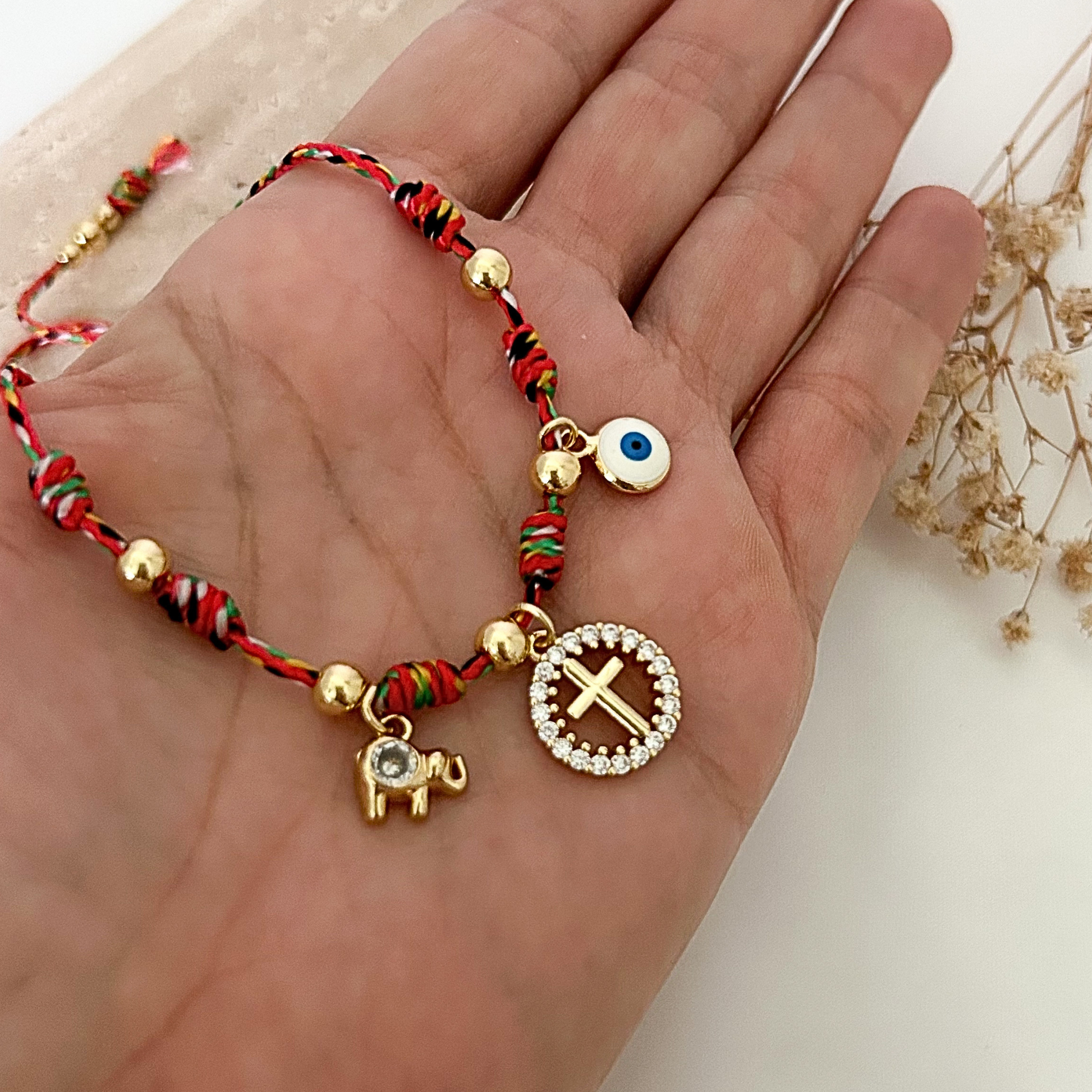 Close-up of a colorful adjustable bracelet inspired by faith and protection, featuring a rhinestone cross charm, evil eye charm, and gold elephant pendant against a soft neutral background.