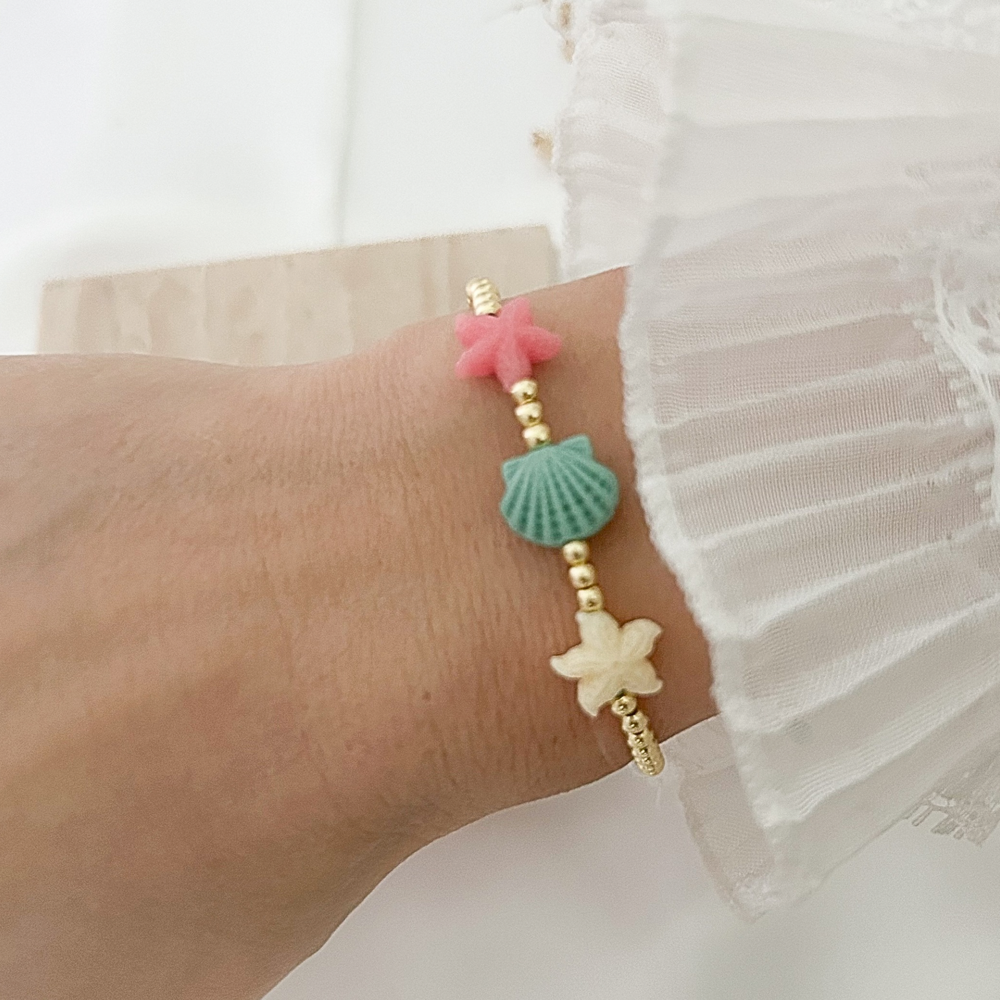 Delicate coastal-inspired bracelet worn on the wrist, designed with gold beads and colorful ocean charms including a pink starfish, aqua seashell, and ivory starfish, paired with a white ruffled sleeve.