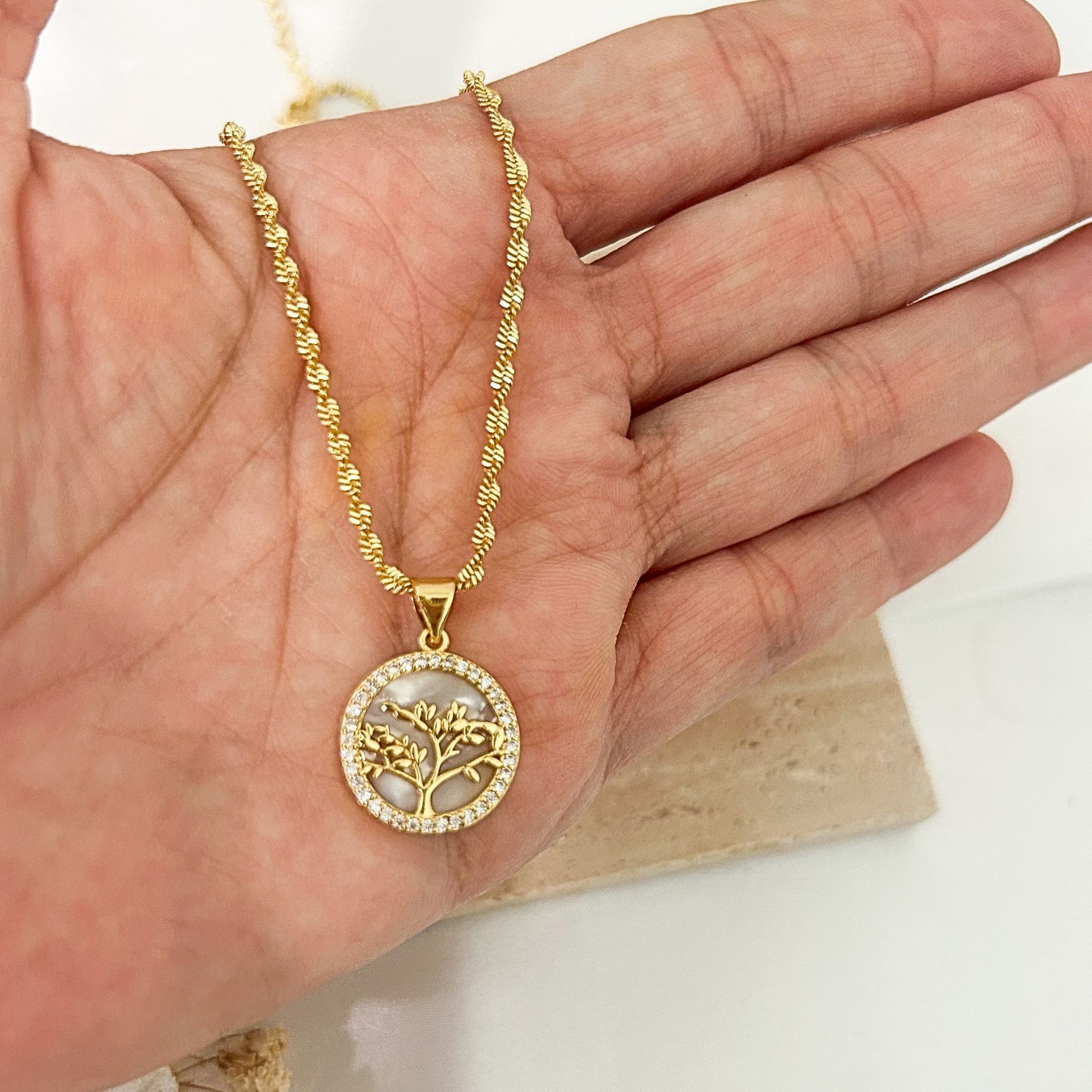 Gold Tree of Life pendant necklace with a mother-of-pearl background and cubic zirconia border, shown in hand to highlight its size, elegant details, and textured gold chain.