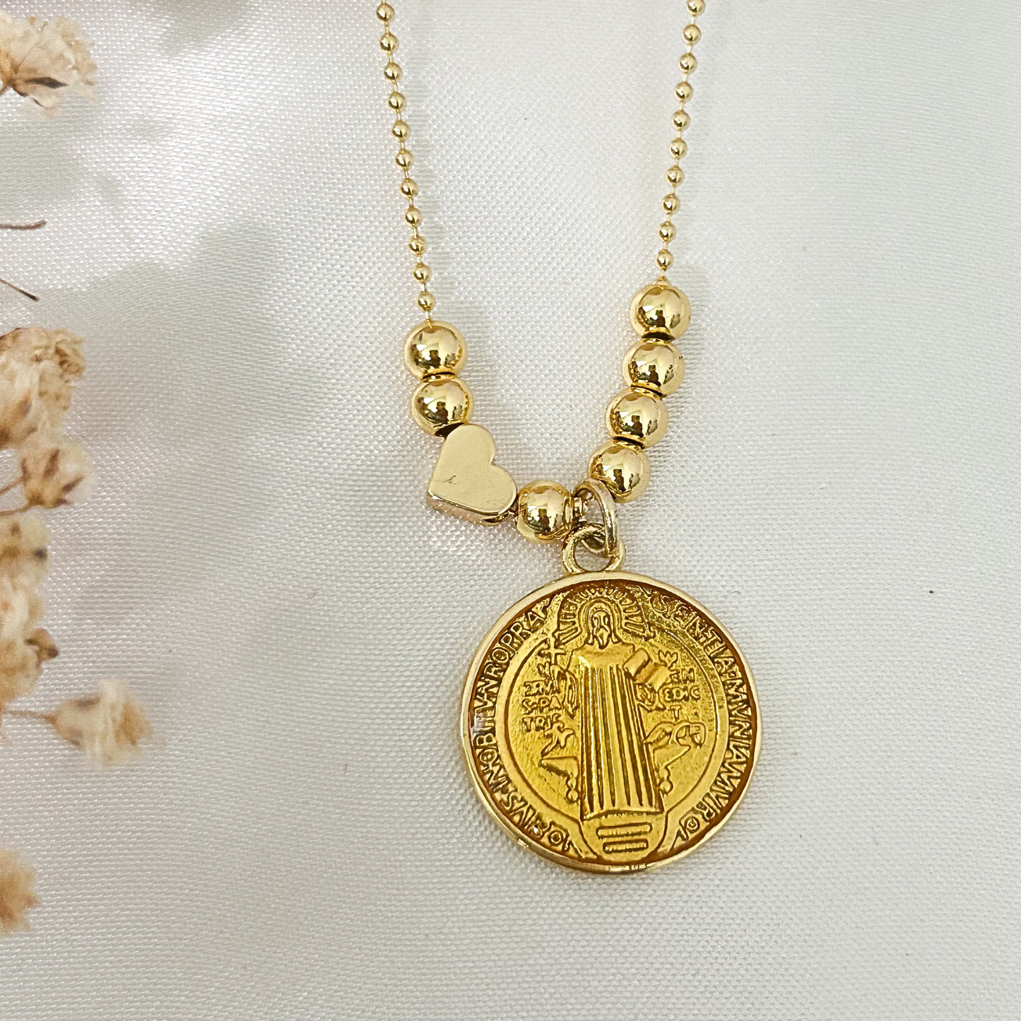 Close-up of a gold Saint Benedict medallion necklace featuring polished gold bead accents on a delicate ball chain, photographed on a neutral minimalist background.