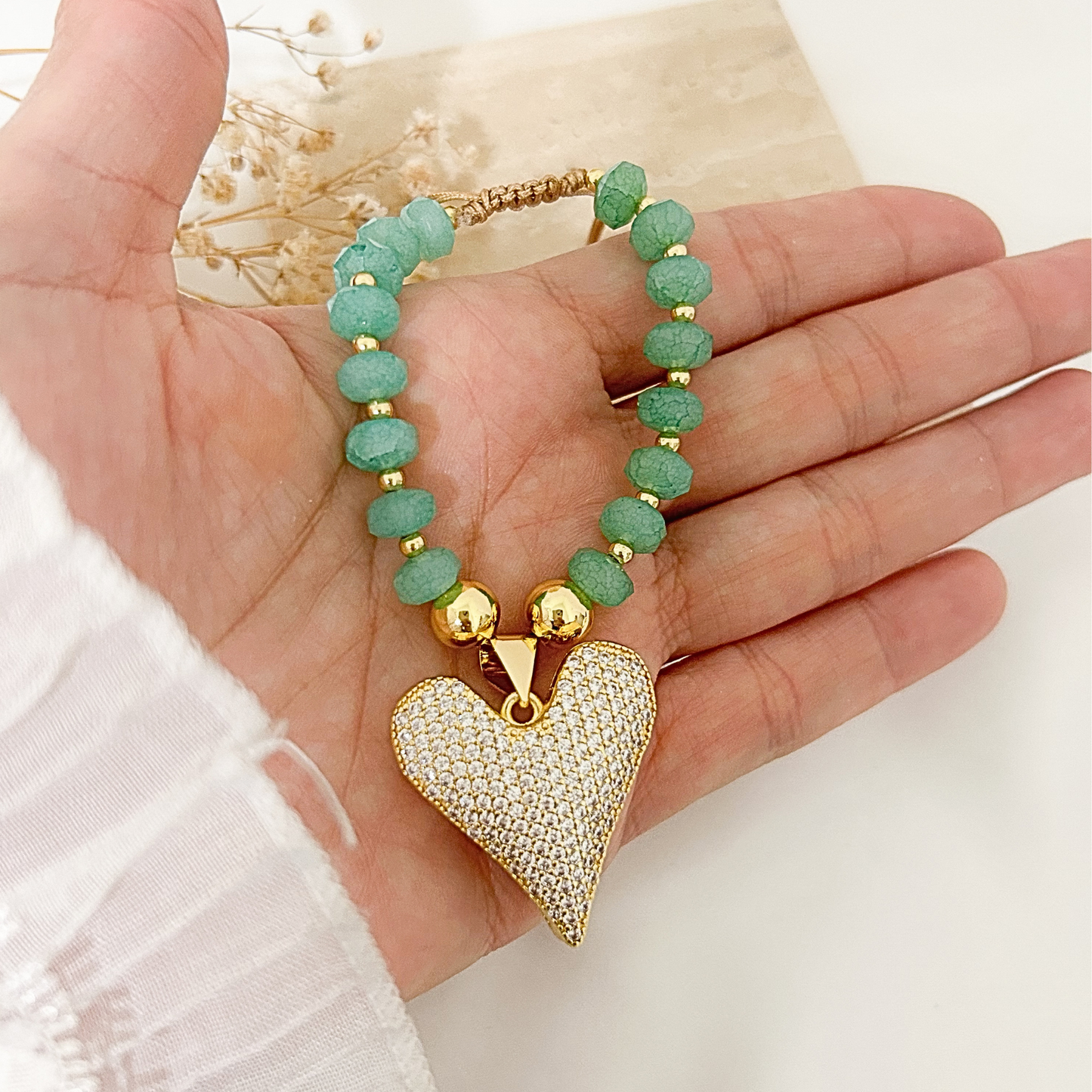 A gold-plated beaded bracelet with textured green beads and a large pave-encrusted heart pendant, held in a person's hand.