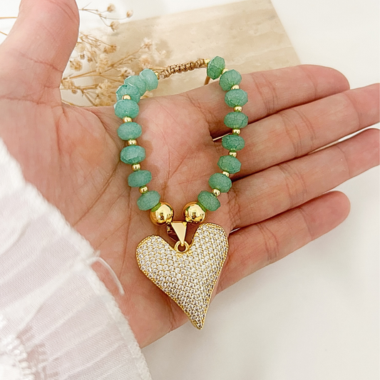 A gold-plated beaded bracelet with textured green beads and a large pave-encrusted heart pendant, held in a person's hand.