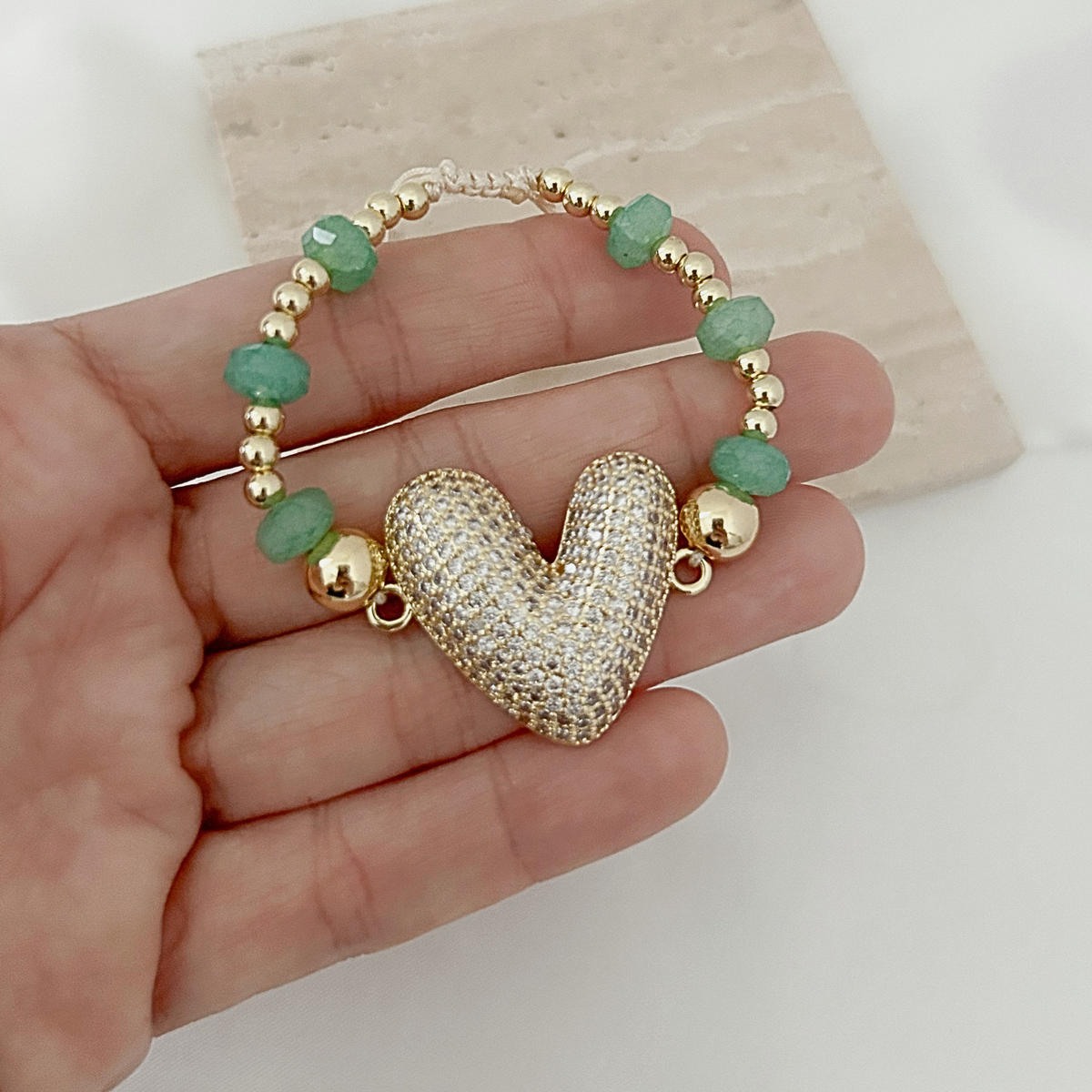 A gold-plated beaded bracelet with a textured heart charm and green faceted beads, held in a hand against a light background.