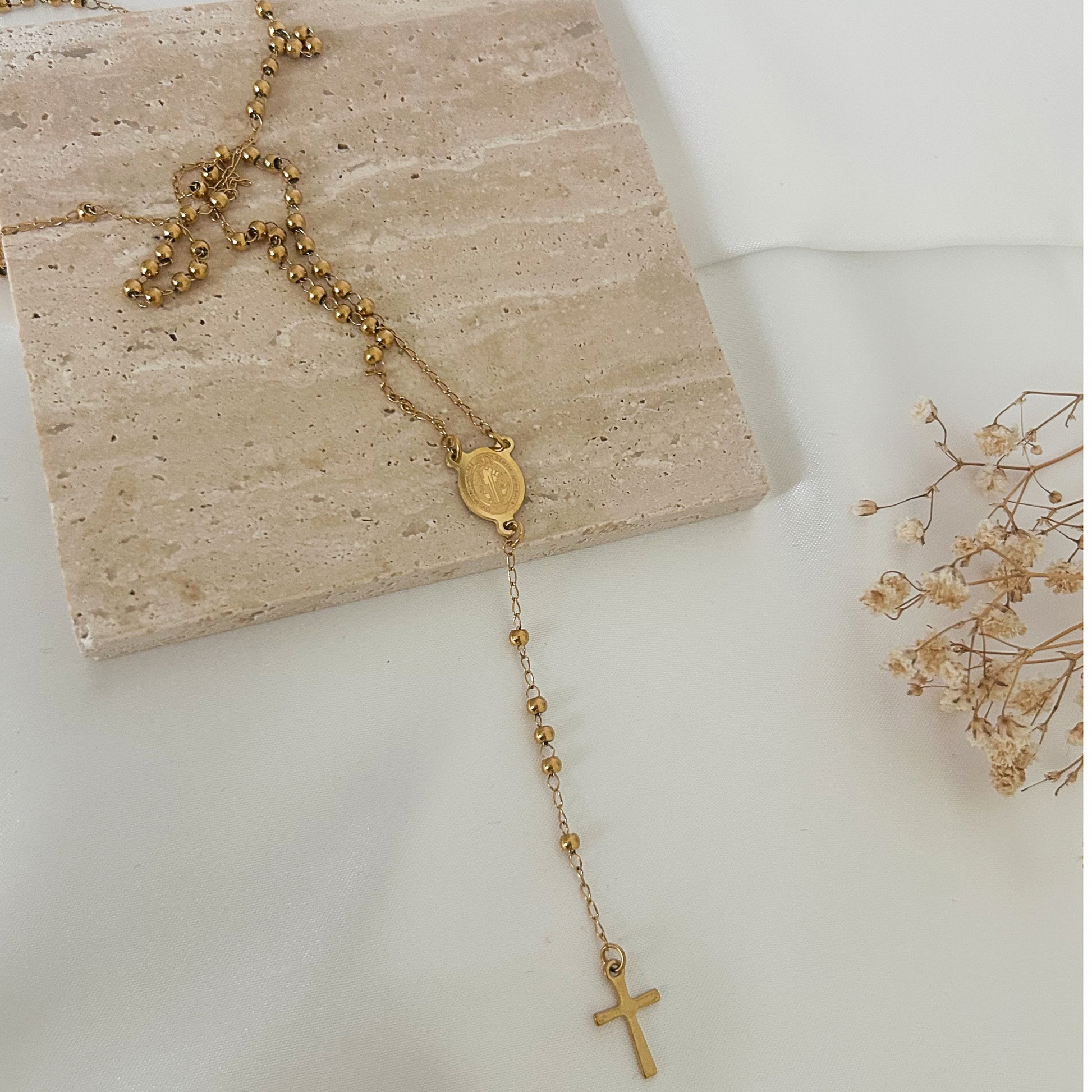 Gold-tone rosary-inspired necklace featuring a Saint Benedict medal centerpiece and a delicate dangling cross, displayed on a neutral stone background with soft floral accents.