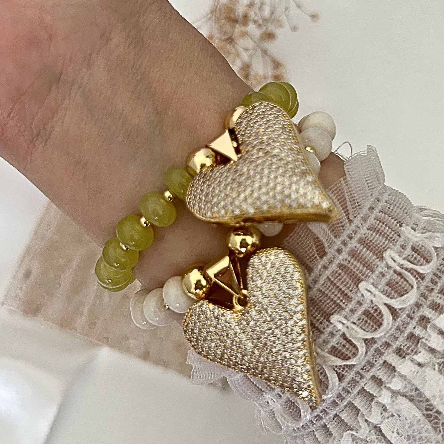 Close-up of two adjustable beaded bracelets featuring sparkling pavé heart charms in gold-tone settings. One bracelet showcases soft ivory beads, while the other features olive green beads, creating an elegant stacked look with feminine lace sleeve details.