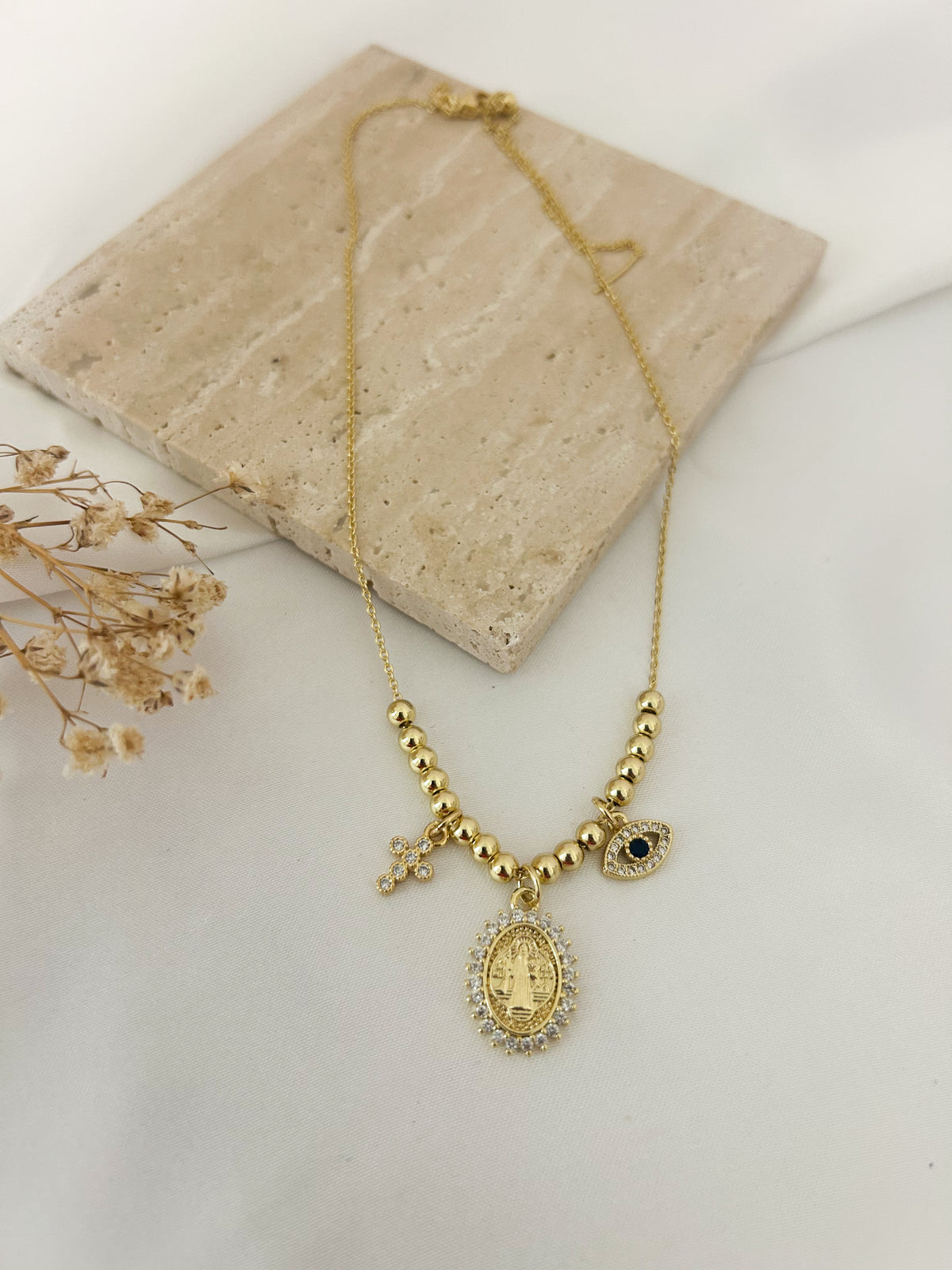 A gold-tone necklace with a central oval religious pendant, small beaded sections, a cross charm, and a small heart charm, displayed on a light beige stone tile with dried flowers.