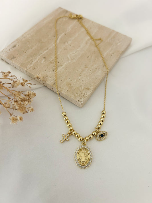 A gold-tone necklace with a central oval religious pendant, small beaded sections, a cross charm, and a small heart charm, displayed on a light beige stone tile with dried flowers.