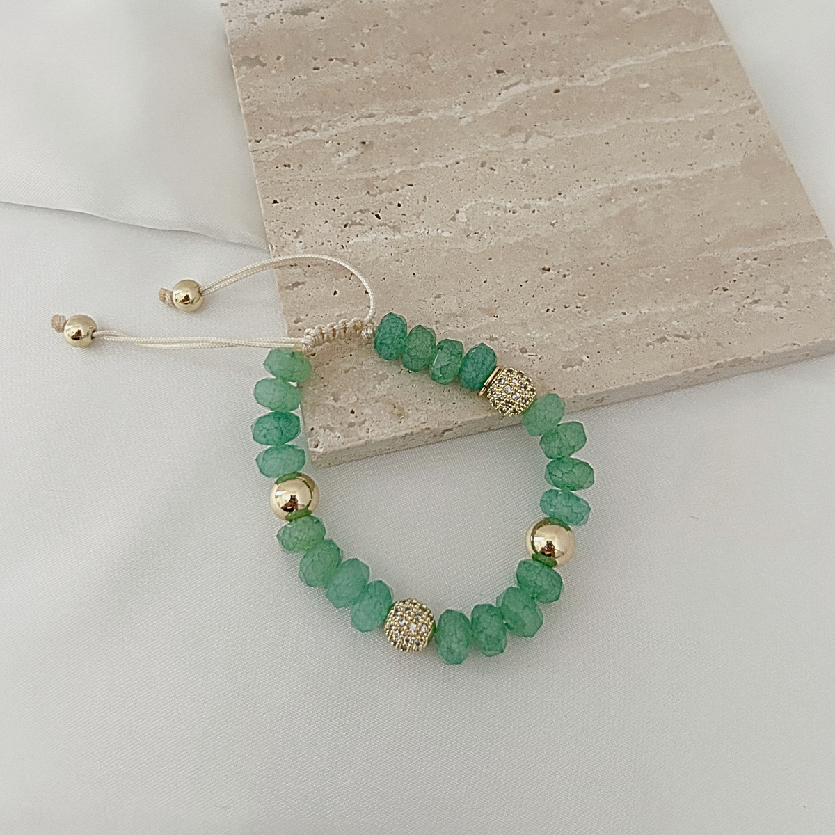 A beaded stretch bracelet made of faceted green beads with gold-toned spacer beads, displayed on a light surface next to a beige stone tile.