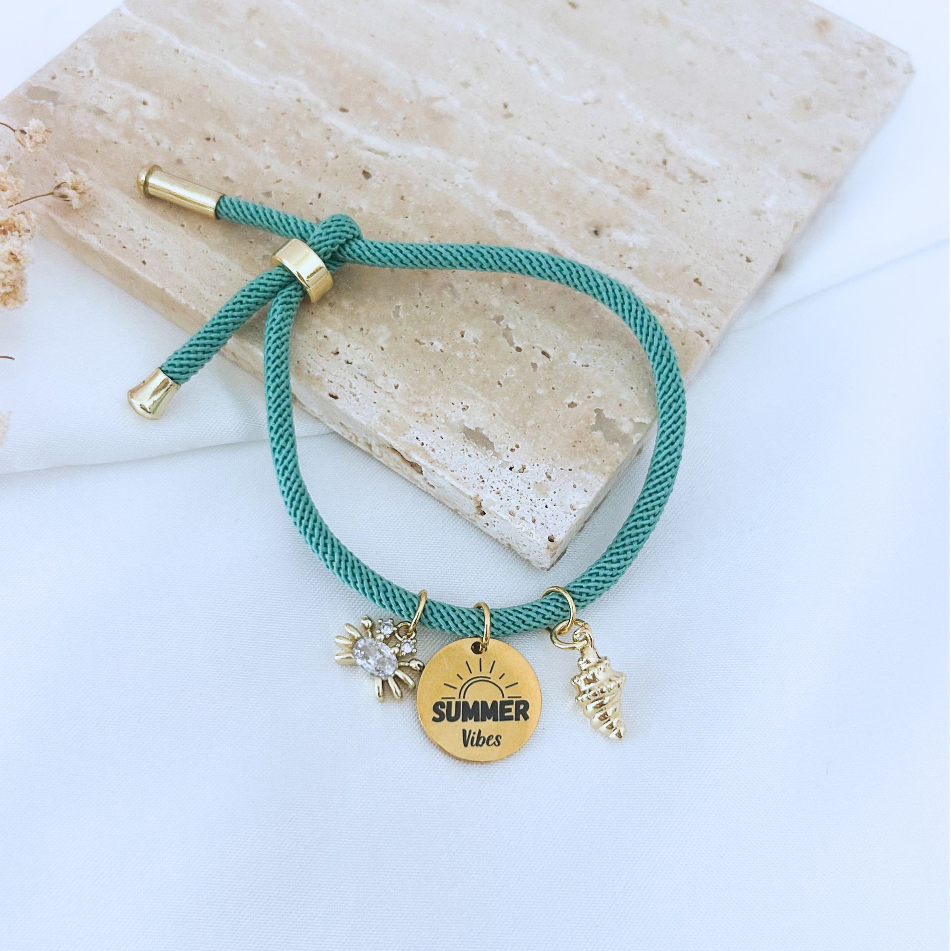 Adjustable teal cord bracelet with gold-tone accents featuring three beach-inspired charms: a sparkling crab, a seashell, and a round “Summer Vibes” charm. Displayed on a neutral stone surface with a soft, minimal background.