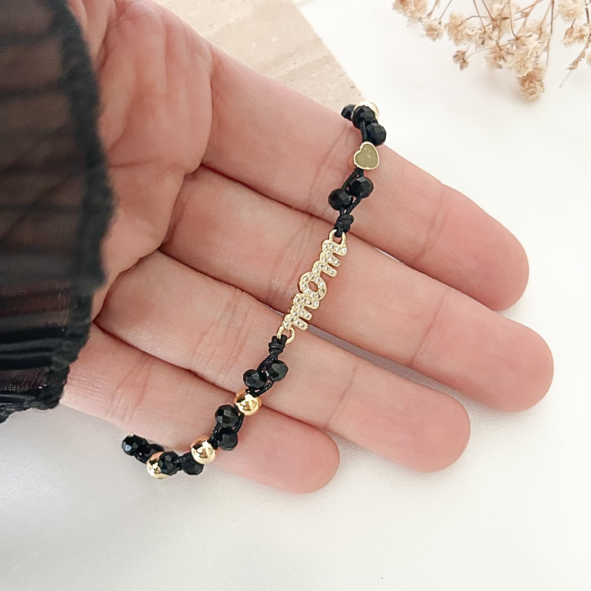 Close-up of a handcrafted black beaded bracelet featuring a sparkling gold-tone “Mom” centerpiece, accented with faceted black crystals and delicate gold heart details, styled against a soft neutral background.