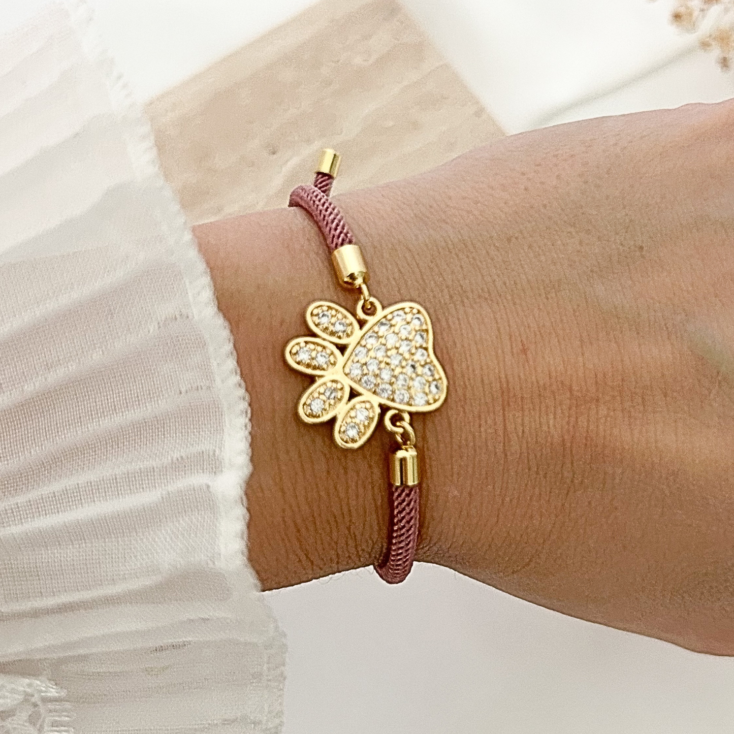 Rose-colored adjustable bracelet worn on the wrist, showcasing a gold paw print centerpiece encrusted with shimmering crystals for an elegant pet-inspired look.