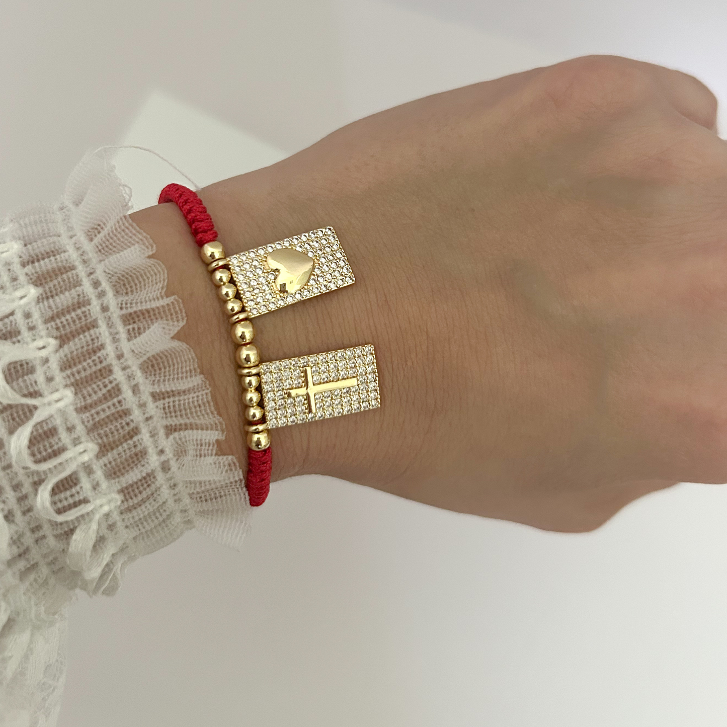 Red cord bracelet with gold-tone cross and heart charms worn on the wrist, highlighting its elegant fit and meaningful design.