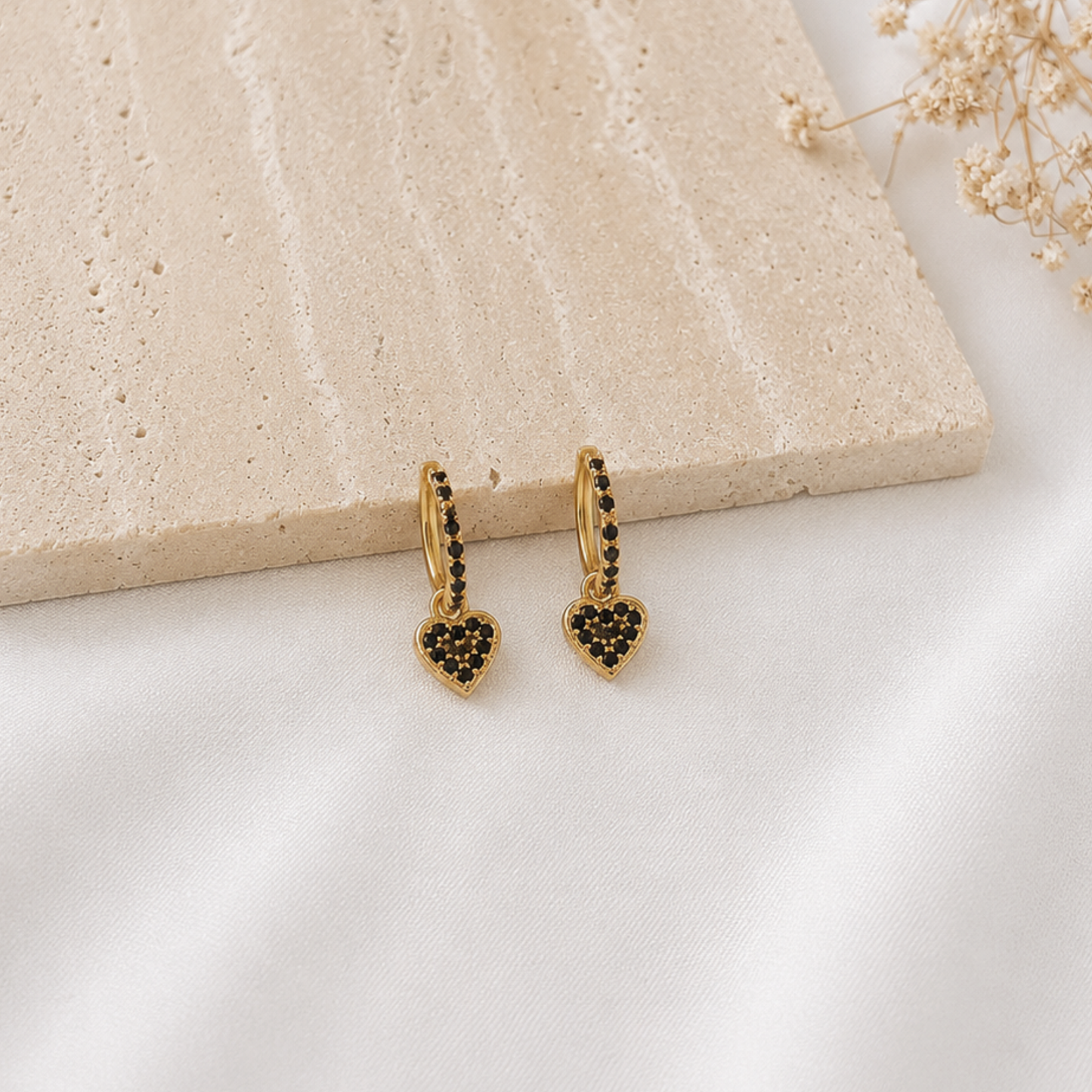 Pair of gold-tone huggie hoop earrings with black pavé crystal detailing and dangling black heart charms. Modern heart earrings with a bold yet elegant design, perfect for everyday wear, date nights, or layering with other gold jewelry.