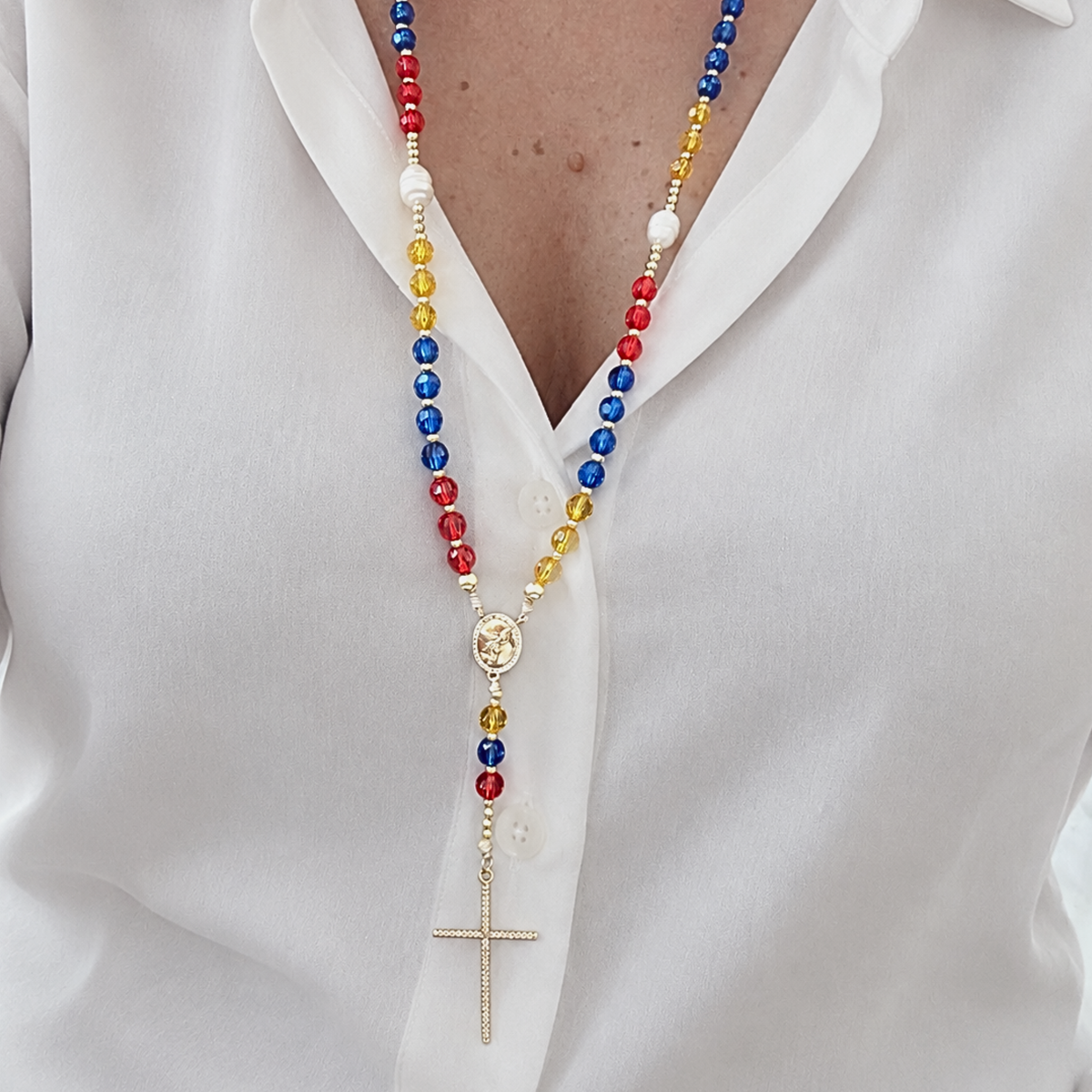 A close-up of a person wearing a long beaded necklace with red, blue, yellow, and white beads, gold-toned links, a round medallion, and a dangling cross pendant over a white collared shirt.