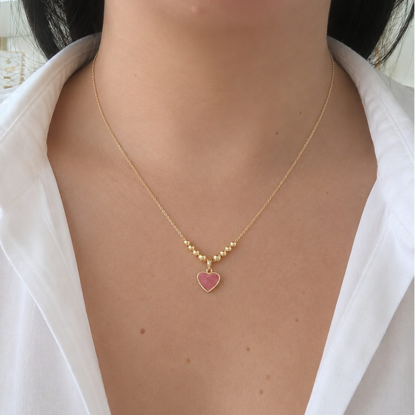 Delicate gold-tone necklace featuring a pink heart pendant accented with polished gold beads, displayed on a neutral stone backdrop with soft dried florals. A feminine piece perfect for everyday wear.