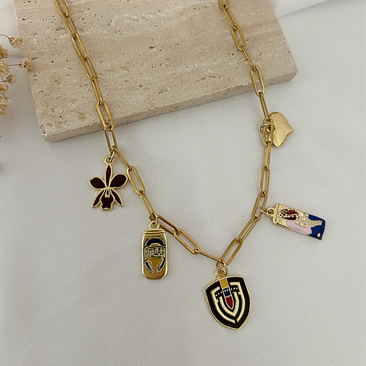 Gold paperclip chain necklace featuring Venezuela-inspired charms, including an orchid, Polar beer can, Savoy chocolate bar, Venezuelan shield, and heart charm, displayed on a neutral minimalist background.
